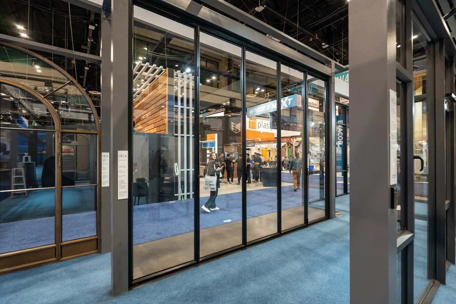 View through glass doors into an exhibition hall with people walking and displays, including signs for 'PURSUIT OF A NEW' and 'EME'