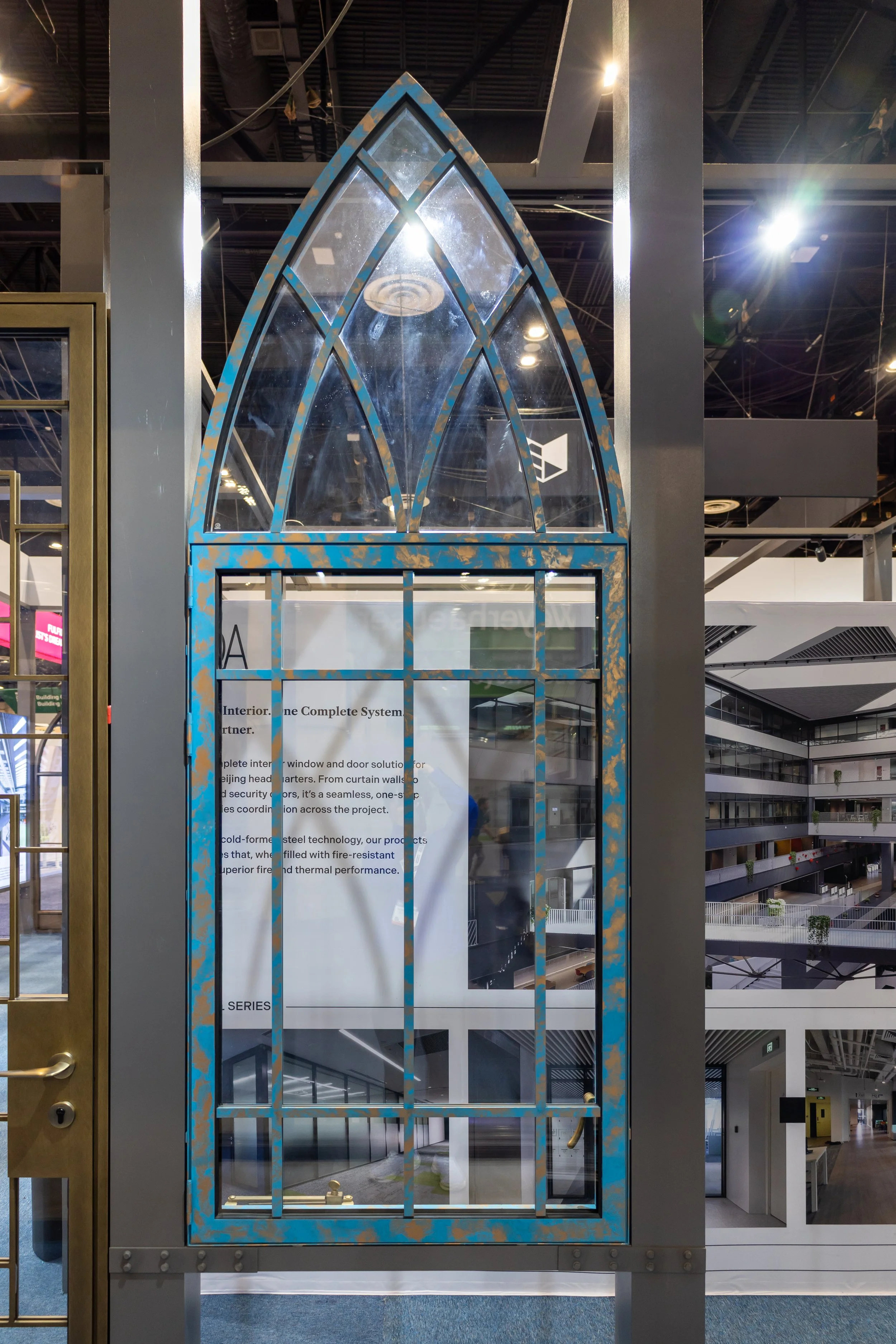 A decorative architectural window with a pointed arch design, made of glass and metal, displayed indoors with a partial view of informational posters and an atrium in the background.