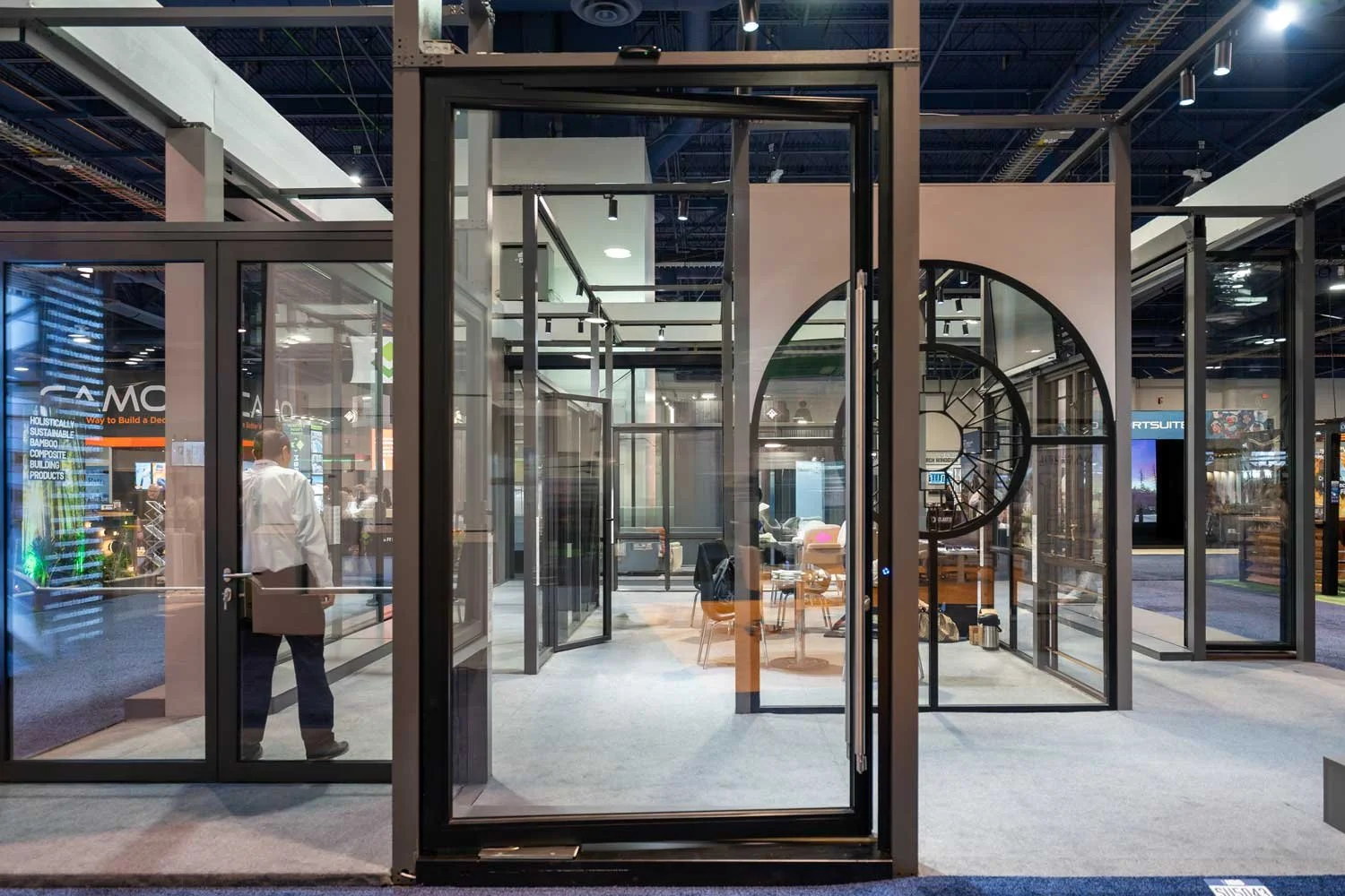 Modern glass-walled office or exhibit space with geometric black frames, featuring interior furniture including chairs and tables, in a professional or trade show environment.