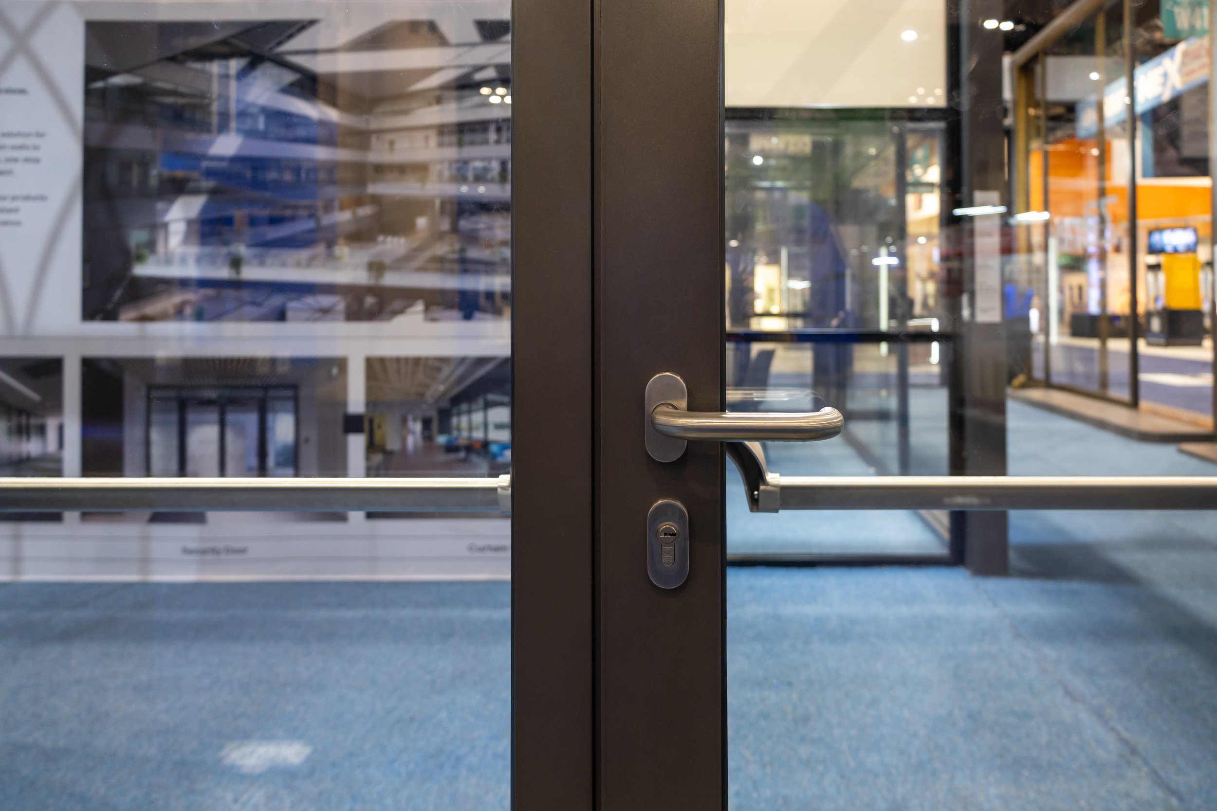 A closed glass double door with a metal handle and lock in an indoor public space, with reflections of the interior and exterior visible on the glass.