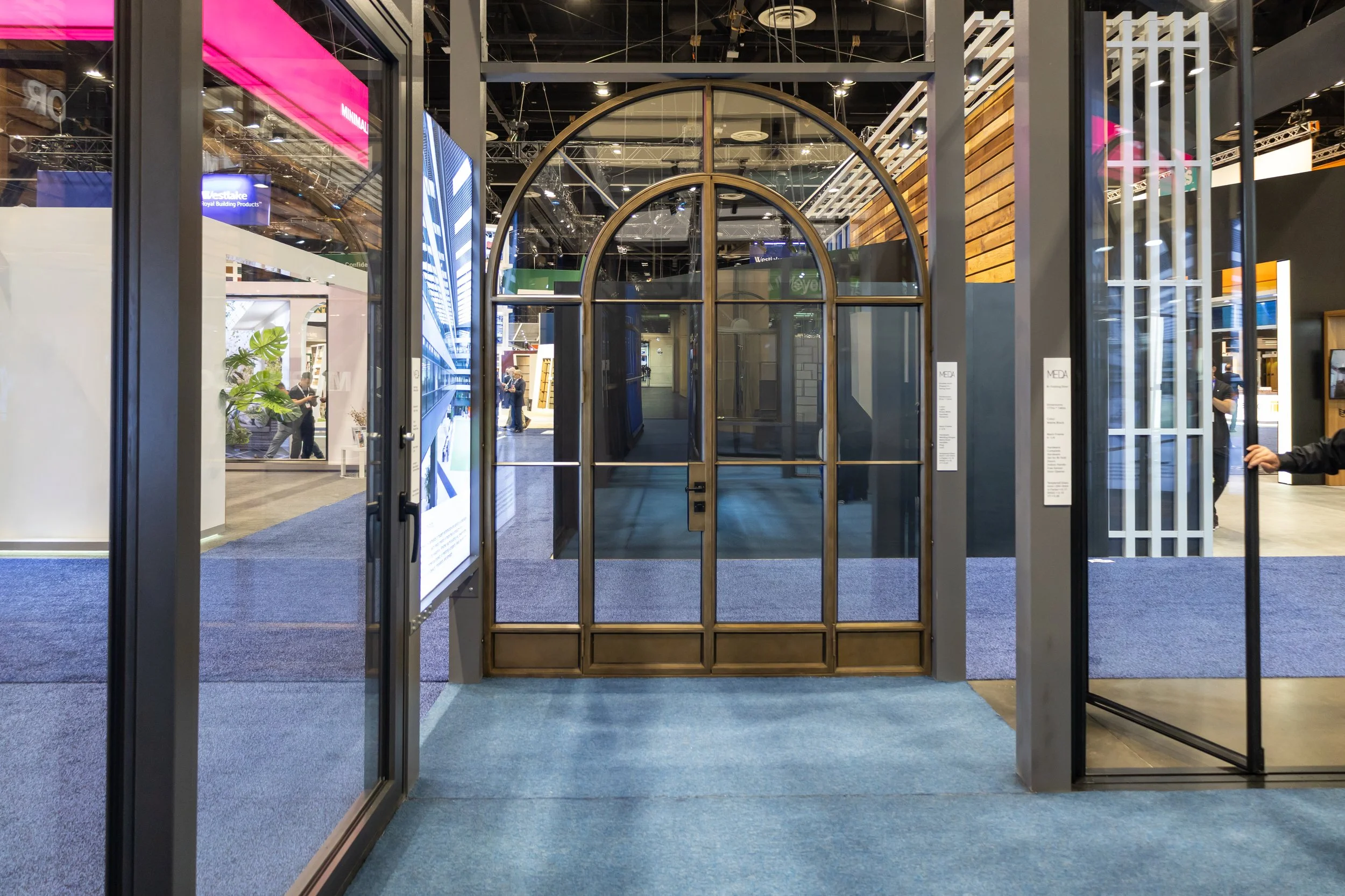 A glass and metal framed door with an arched top leading into a modern exhibition space at a trade show or convention, with other booths and displays visible in the background.