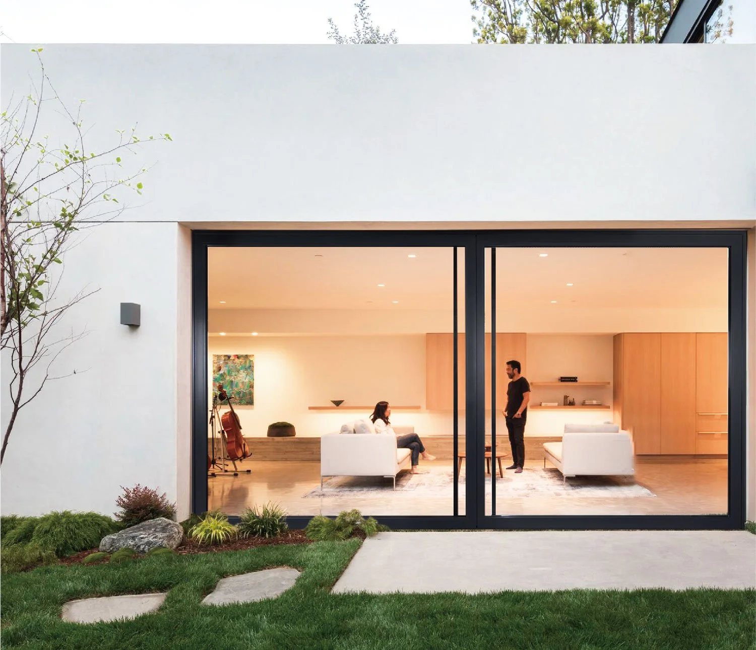 Modern house with large glass sliding doors revealing a living room with two people inside, surrounded by a well-maintained garden with rocks and bushes.