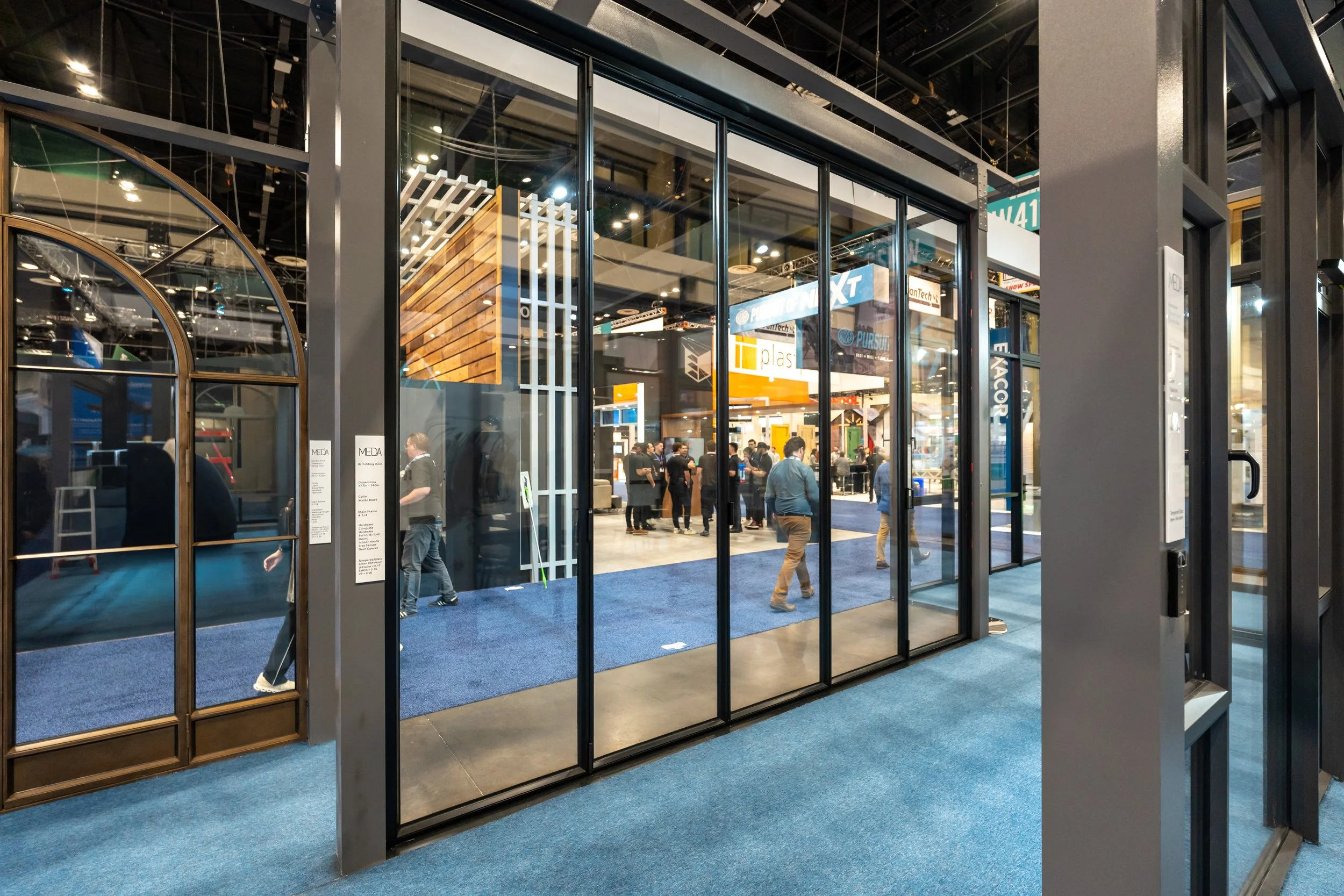 View of an exhibition hall from outside a glass door, showing people walking and displays inside with bright banners and signs.