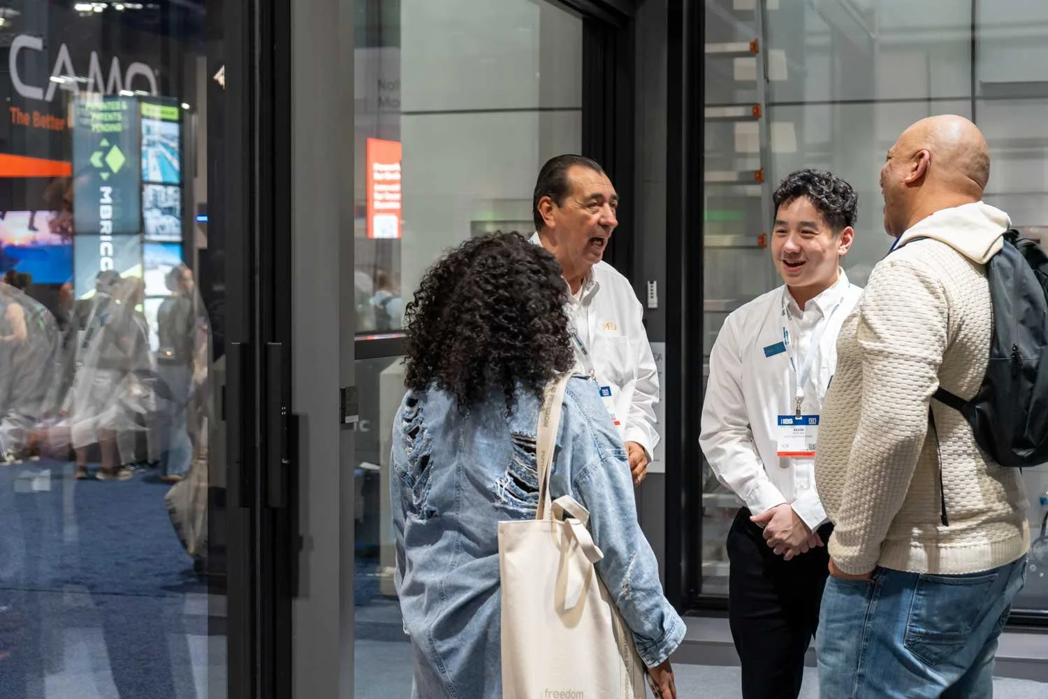 People talking in an indoor setting, likely at a conference or event, with a glass door behind them reflecting other attendees.