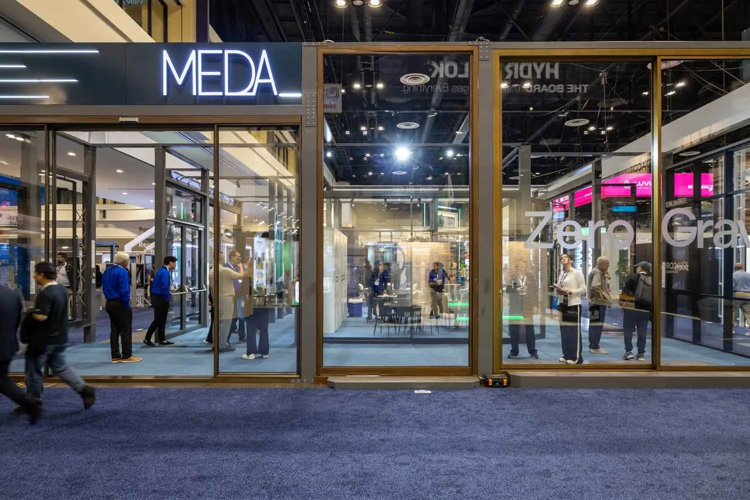A trade show booth with glass walls, labeled 'MEDA' at the top left corner and 'Zero-Gravity' on the right side, with people inside and outside the booth.