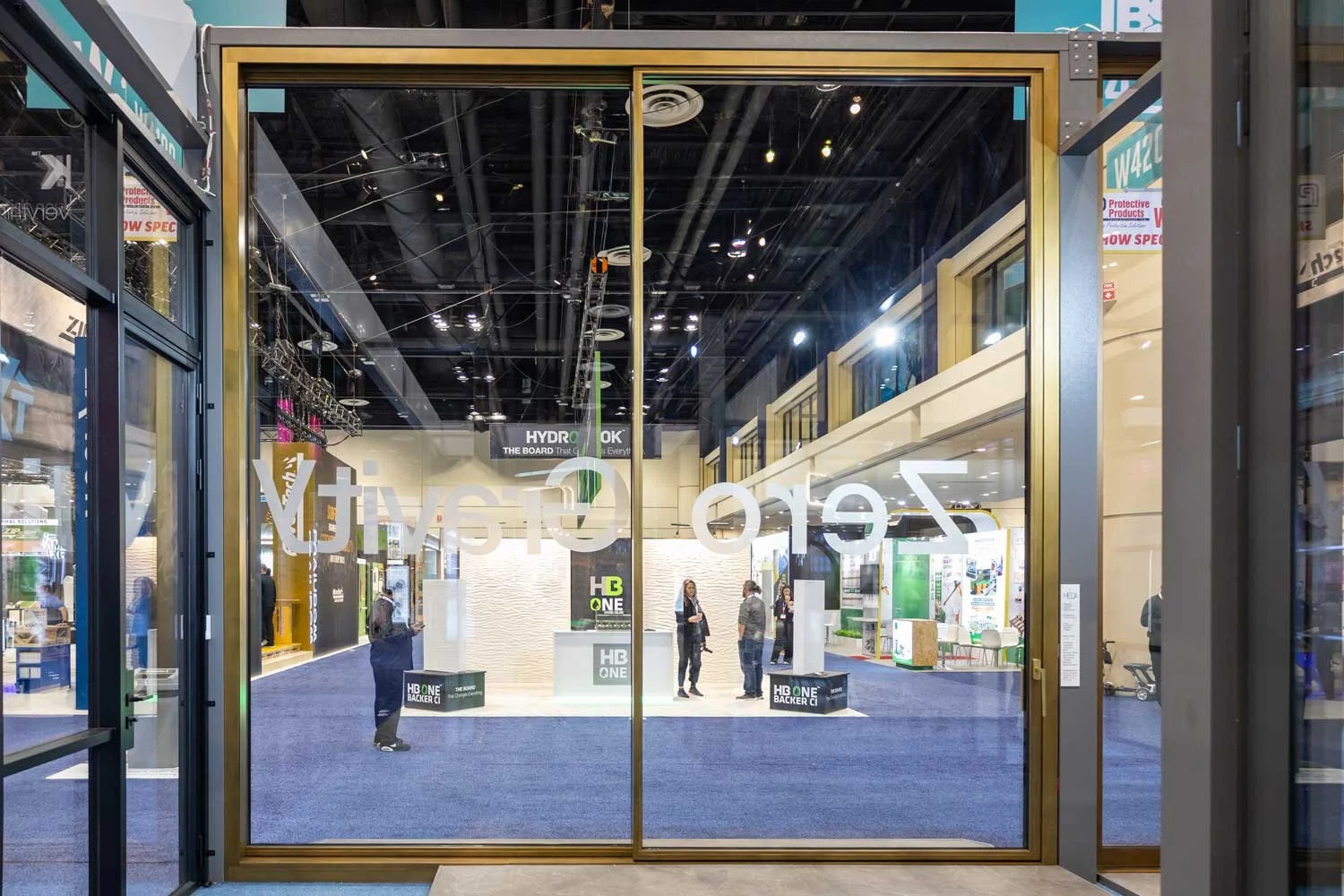 View of a trade show or conference exhibition inside a large hall, seen through glass doors with gold-colored frames. Several attendees are present, some talking and others walking, with booths and displays in the background.