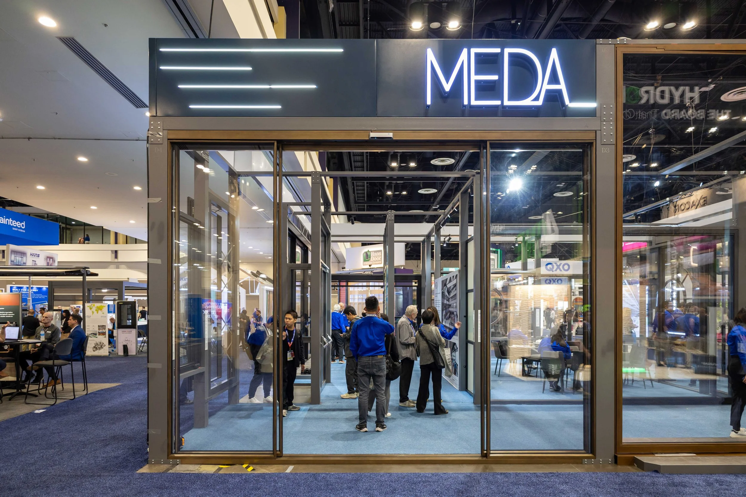 A trade show booth with the name 'MEDA' illuminated in blue neon lights on a black background. Inside the booth, people are engaged in conversations and viewing displays.