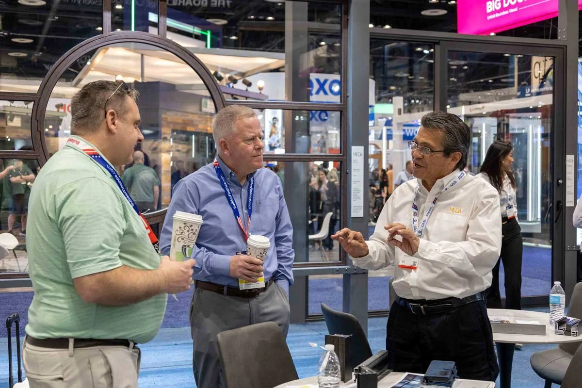 Three men in business casual attire are having a conversation in a trade show booth, holding coffee cups. The man on the right is gesturing with his hands while talking. Other people and booths are visible in the background.