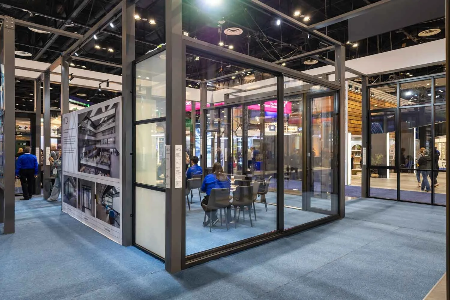 A clear glass-walled booth at a trade show with people sitting inside and nearby, with displays and signage in the background.