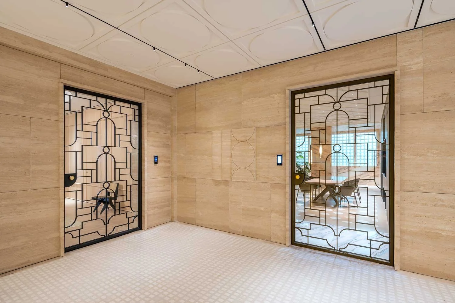 Interior of a modern building with two glass doors featuring black geometric patterns, beige stone walls, a light-colored step patterned floor, and a view into a room with a dining table and chairs in the background.