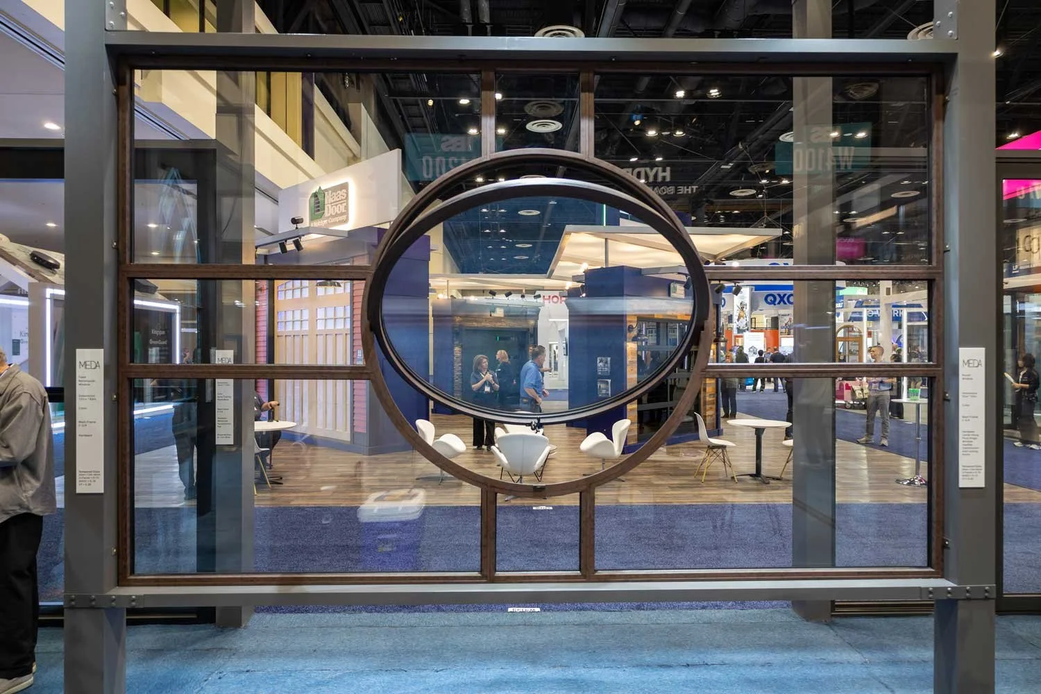 A large glass and metal frame display with a circular mirror inside, showing a busy exhibition hall with several booths and people.