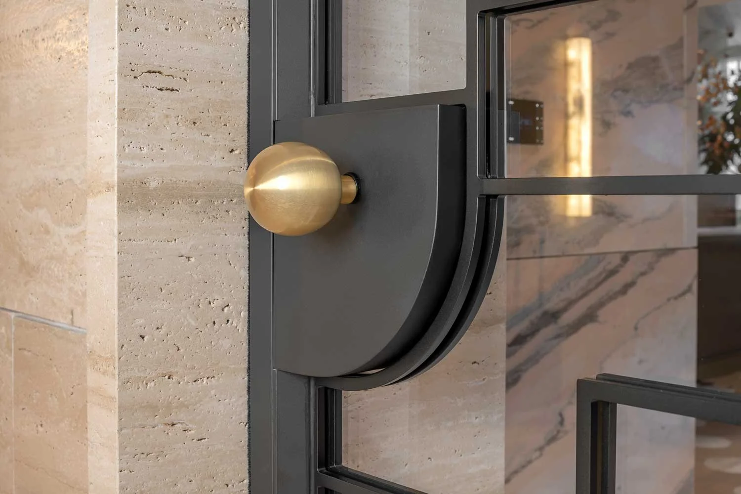 Close-up of a decorative black metal and gold door handle with a round gold knob on a black curved plate, mounted on a beige stone wall.