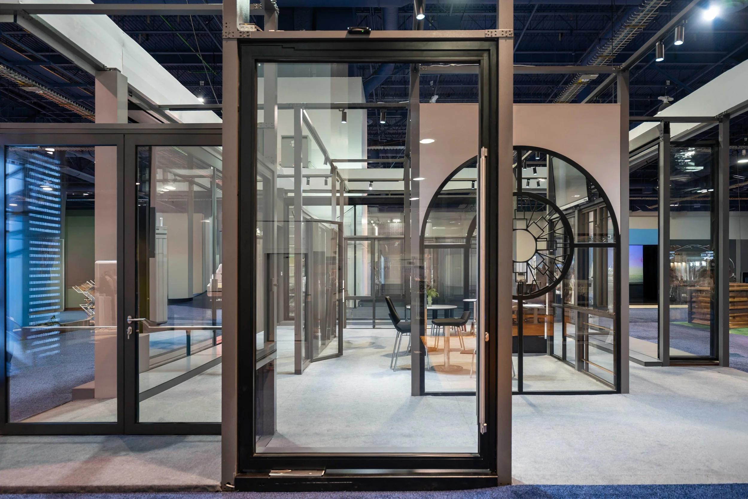 Interior view of a modern glass-walled office space or meeting room with visible chairs and tables, inside a large exhibition or showroom with high ceilings and track lighting.
