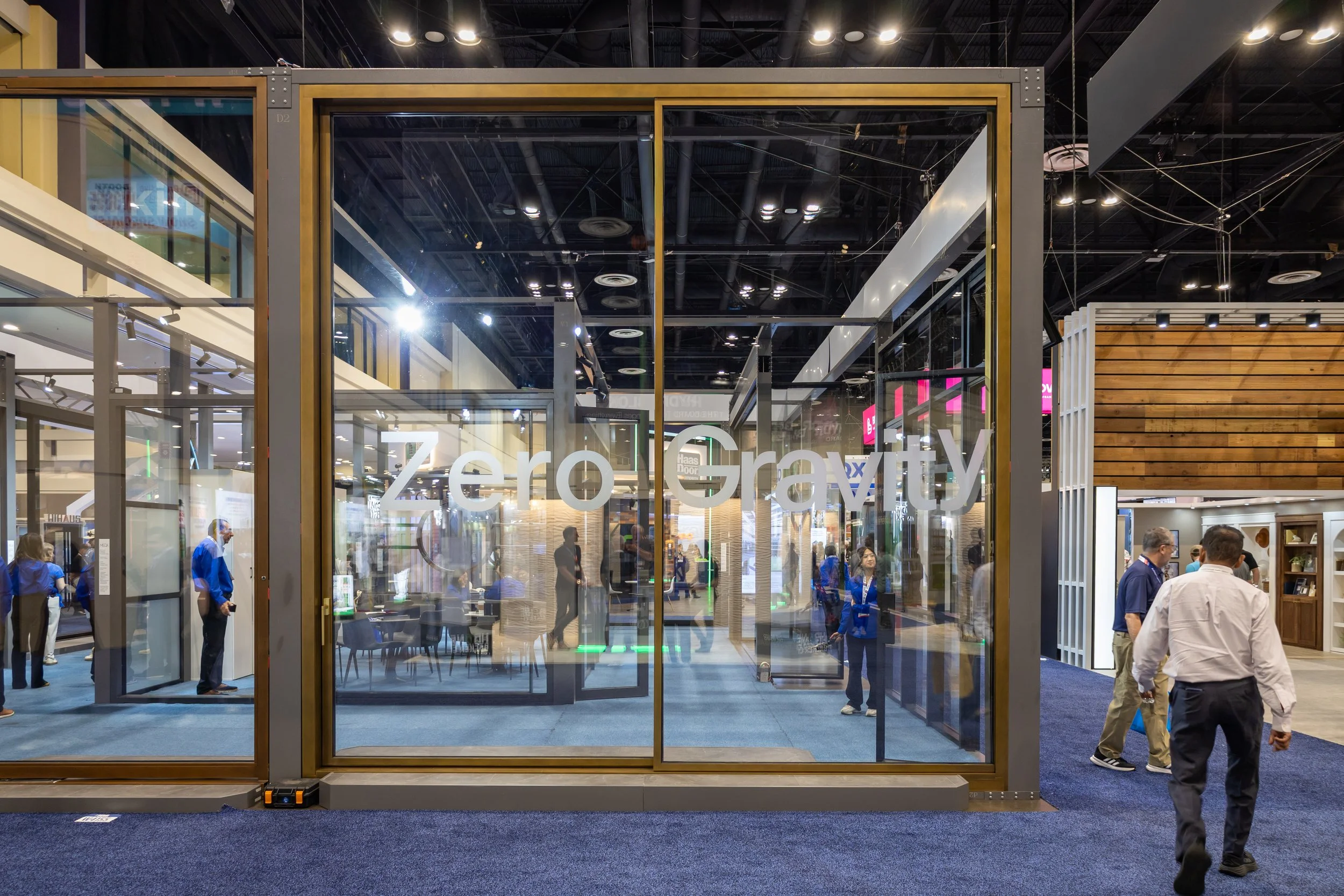 A glass enclosure with a sign that reads 'Zero Gravity' at an indoor trade show or exhibition, with several people walking and observing displays inside and outside during a busy event.