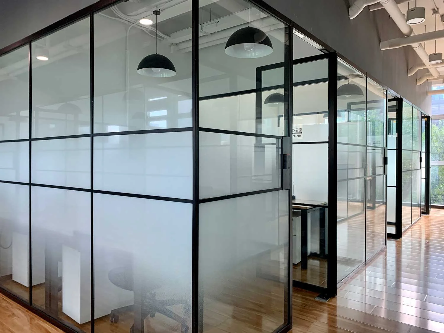Modern glass-walled office cubicles with black framing, pendant lighting, and wooden flooring.
