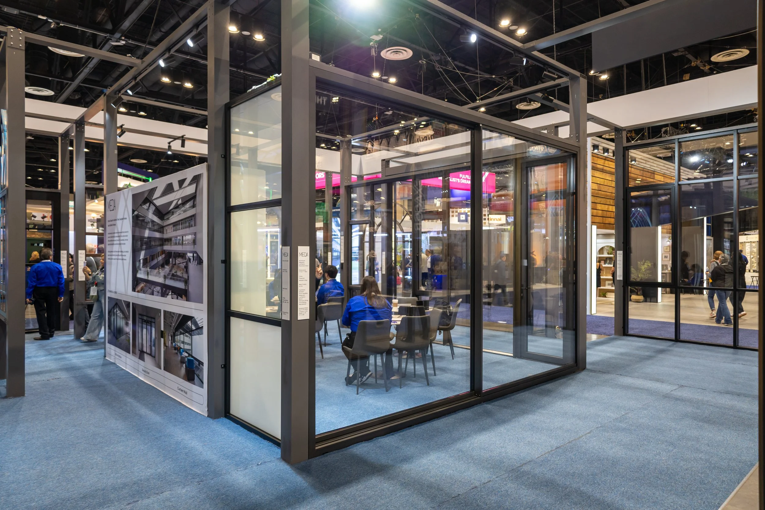A modern exhibition booth with glass walls and a seating area inside at a trade show, with people walking and exploring nearby booths.