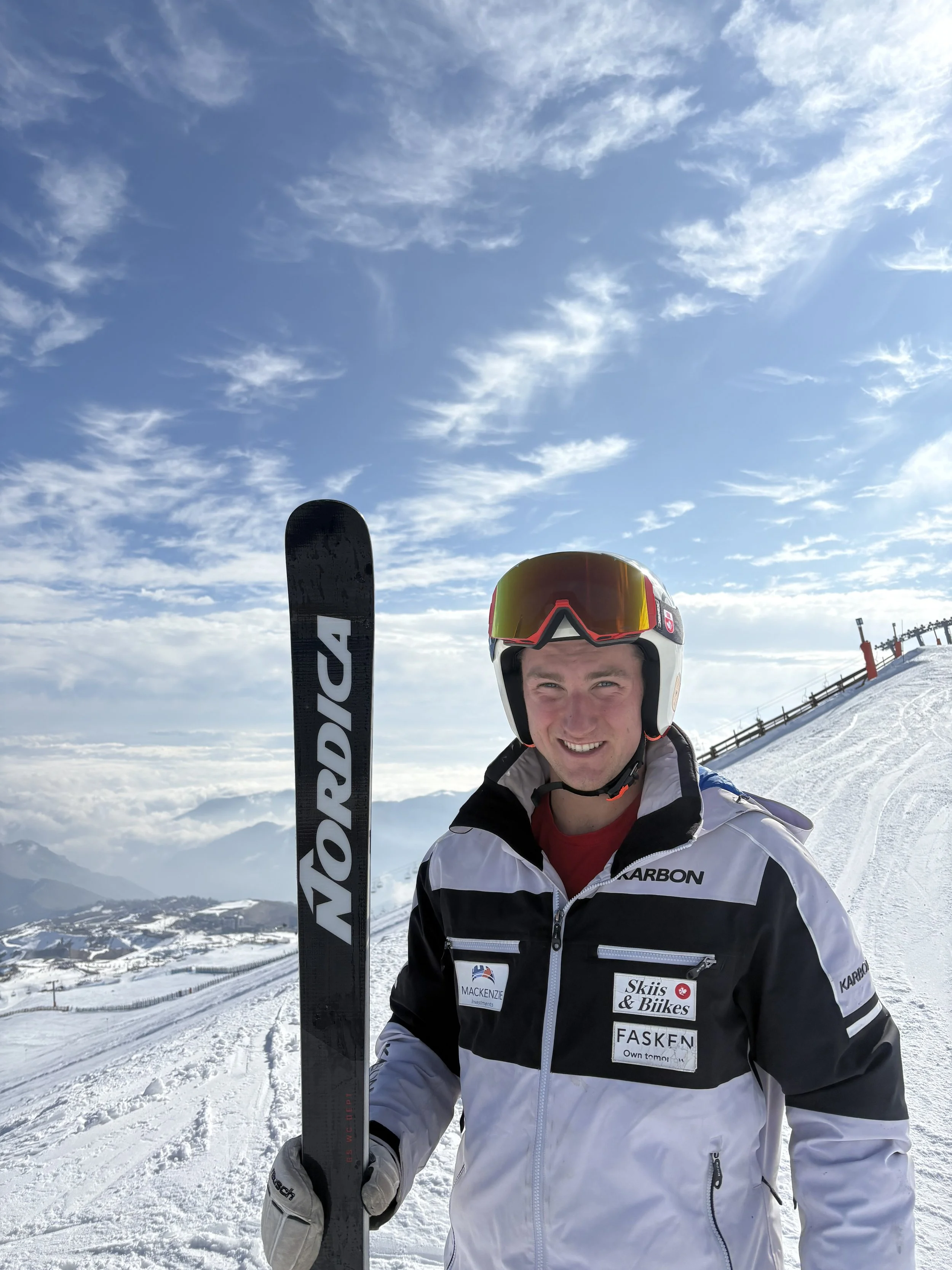 A smiling man in ski gear holding a black Nordica ski on a snowy mountain slope under a partly cloudy sky.
