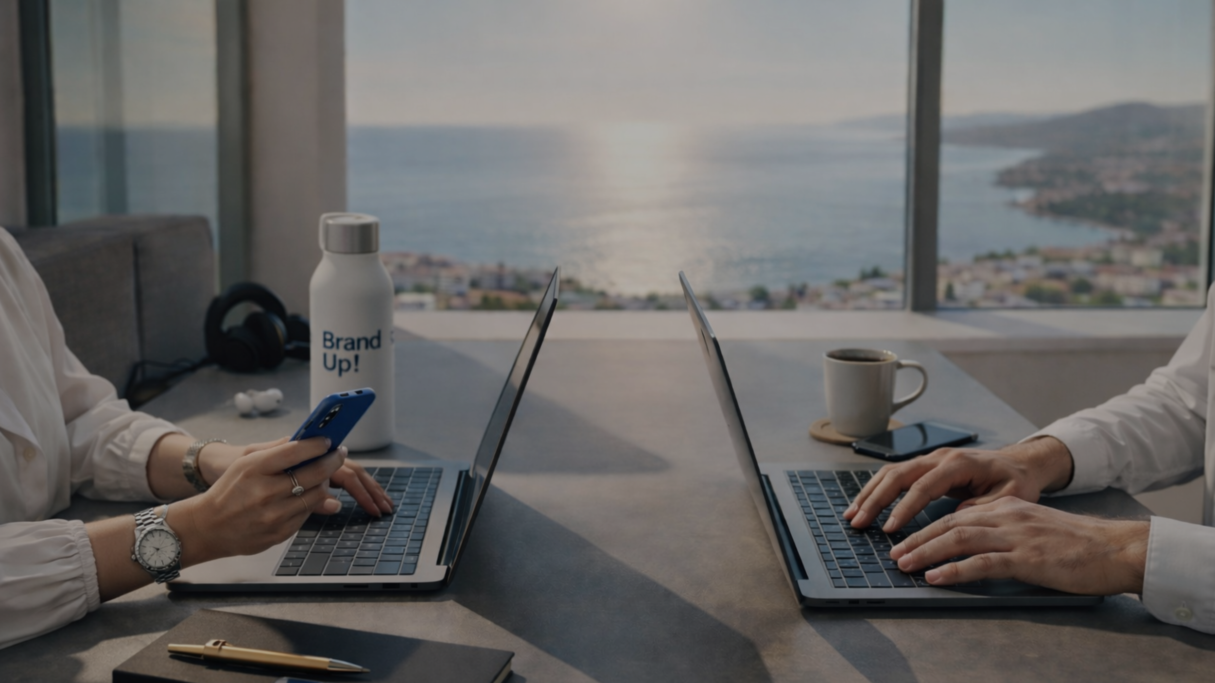 Two people working at a table with laptops, one person holding a smartphone, with a view of water and cityscape outside the window.