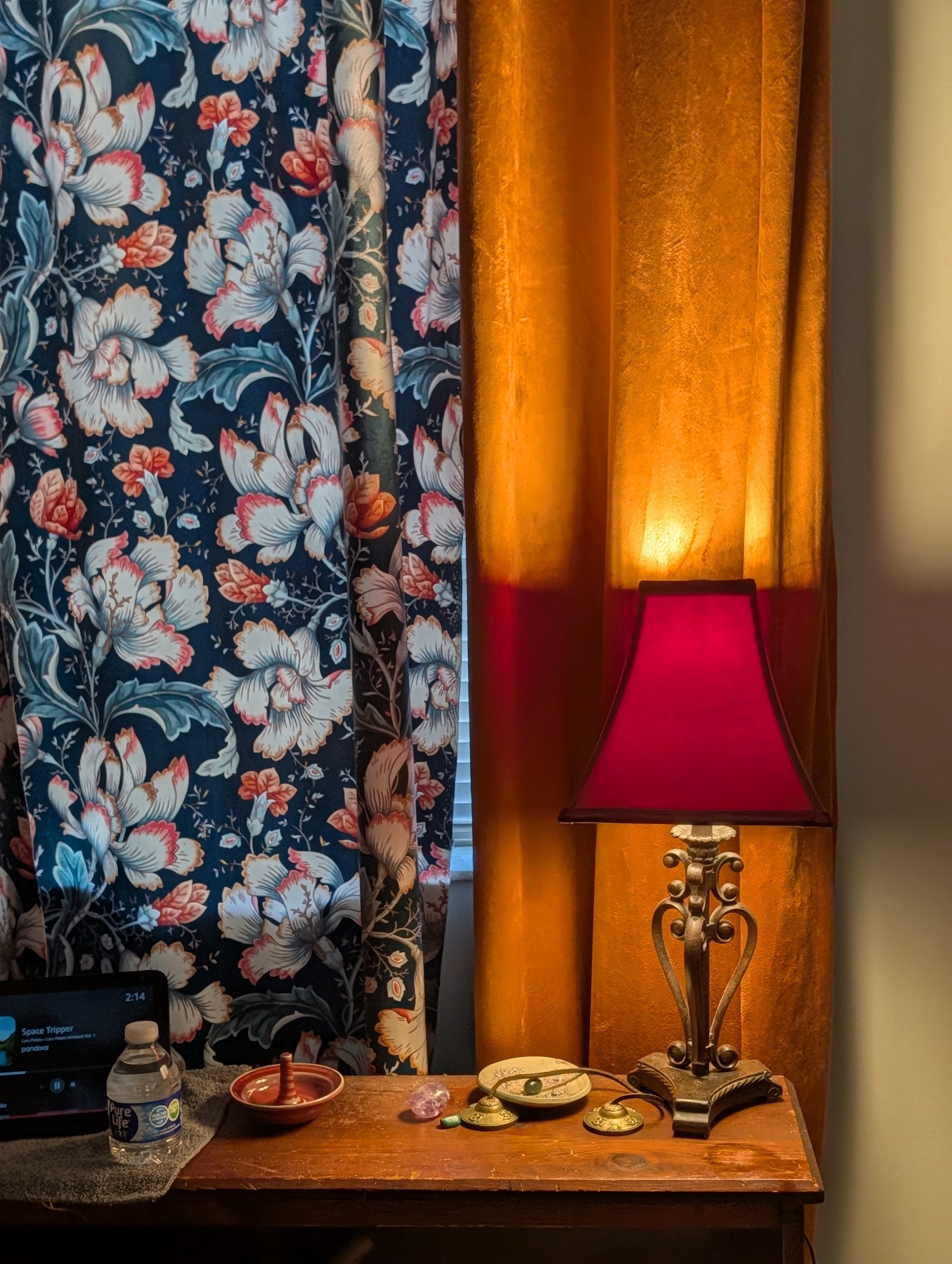 A wooden side table with a bottle of water, a small bowl, a few jewelry pieces, and a tablet playing music, next to a lamp with a red shade. Behind the table, there are floral curtains and orange drapes.