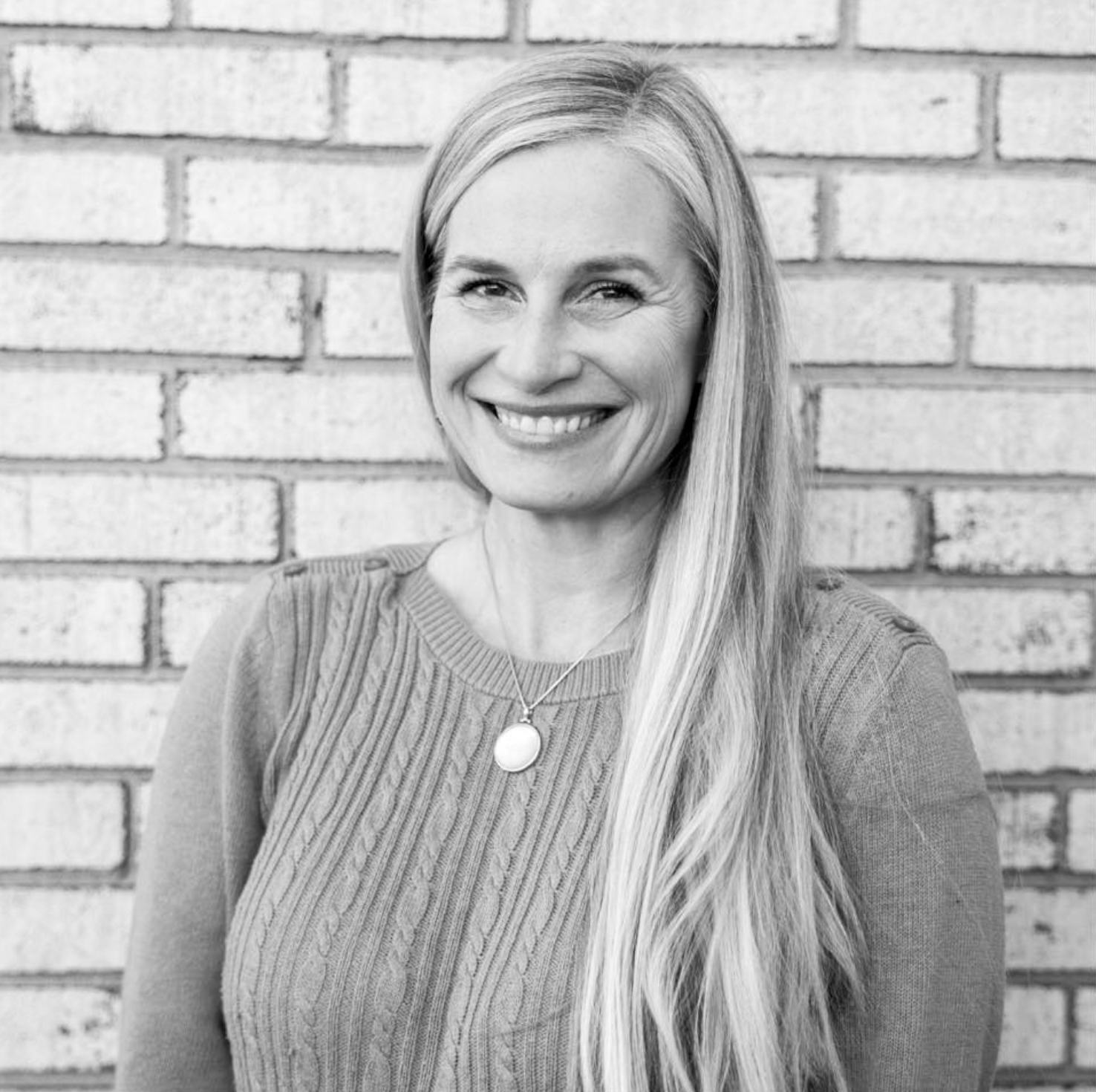Black-and-white portrait of a smiling woman with long blonde hair, wearing a cable-knit sweater and a necklace, standing against a brick wall.