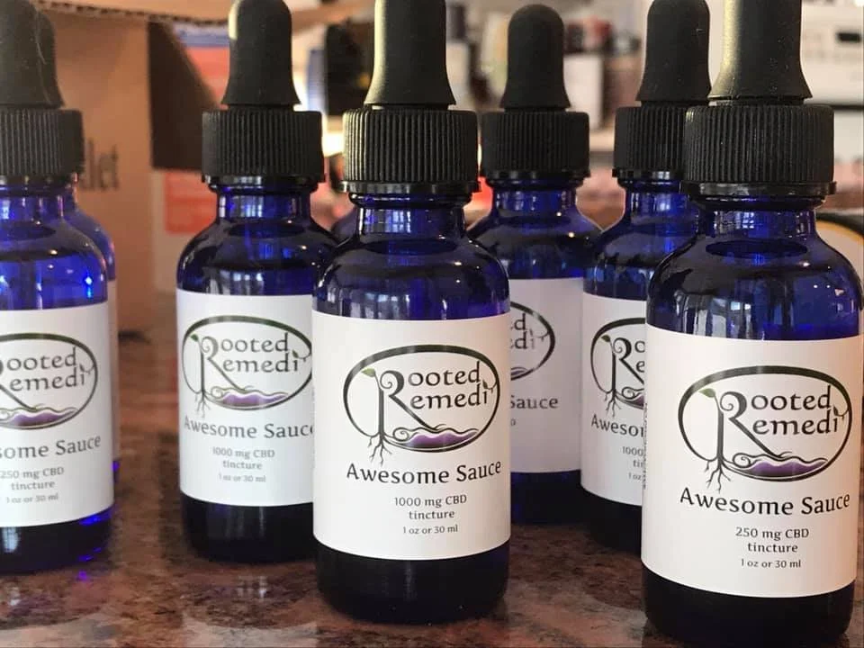 Six blue glass bottles of Rooted Remedi Awesome Sauce CBD tincture with black dropper caps on a countertop.