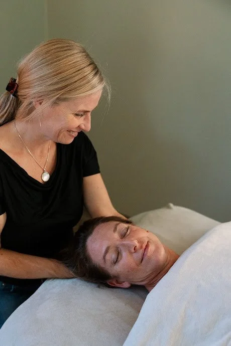 A woman with blonde hair in a ponytail and wearing a black shirt gently offering craniosacral and massage therapy to a woman lying on a massage table who has her eyes closed and appears relaxed.