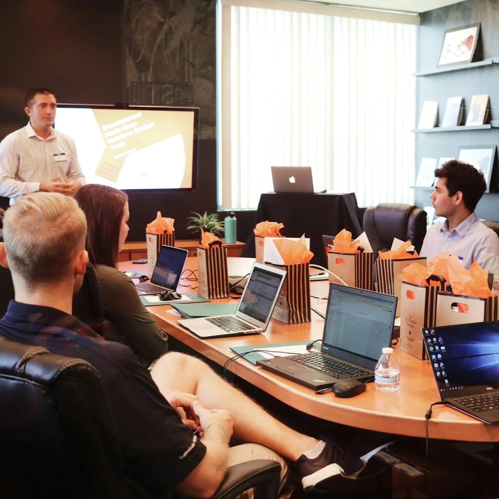 A business meeting or workshop with one person standing near a large screen and five people seated at a conference table with laptops and gift bags, in a modern office setting.