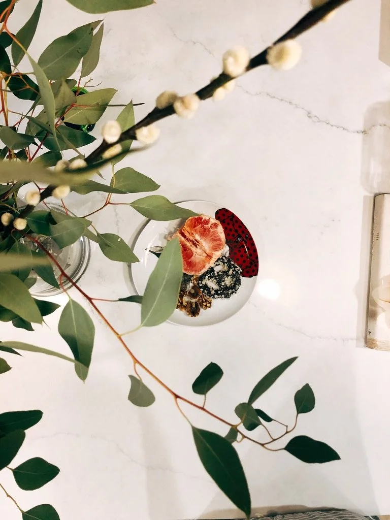 A white marble table with a small white plate holding a peeled tangerine, some dried fruit, and a red-and-black patterned object, partially obscured by green leafy branches.