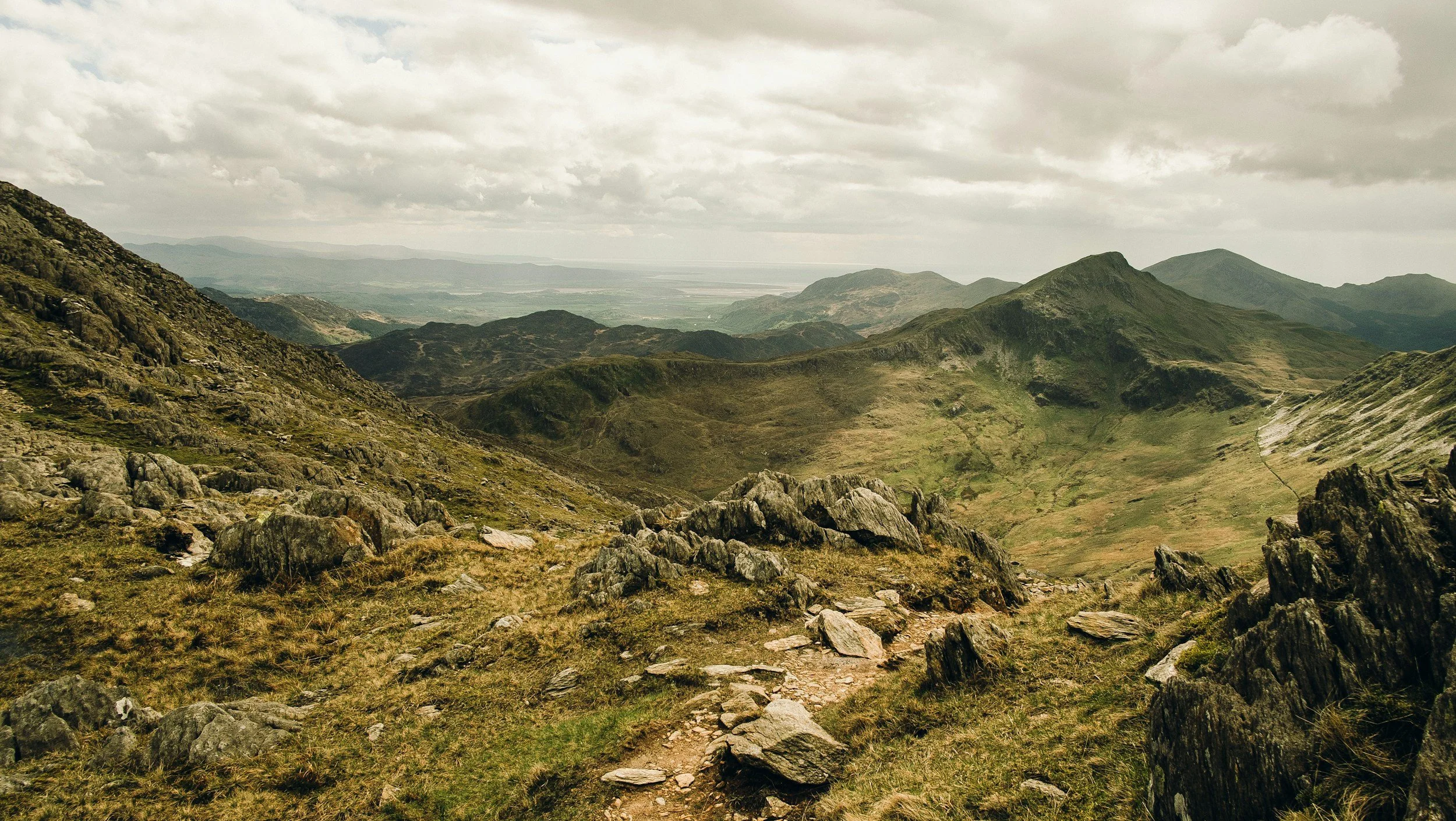 A mountainous landscape with rocky terrain, green grassy slopes, and cloudy sky, possibly a hiking trail.