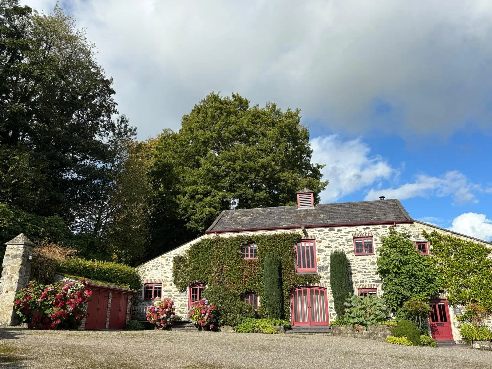 Stone house with pink window frames, surrounded by lush greenery and pink flowers, under a partly cloudy sky.