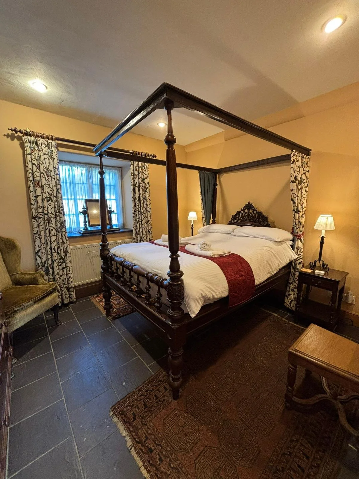 A bedroom with a four-poster bed, ornate headboard, and linen with rolled towels on it, flanked by two lamps. It has dark tile flooring, an armchair, a window with patterned curtains, and a small wooden table.