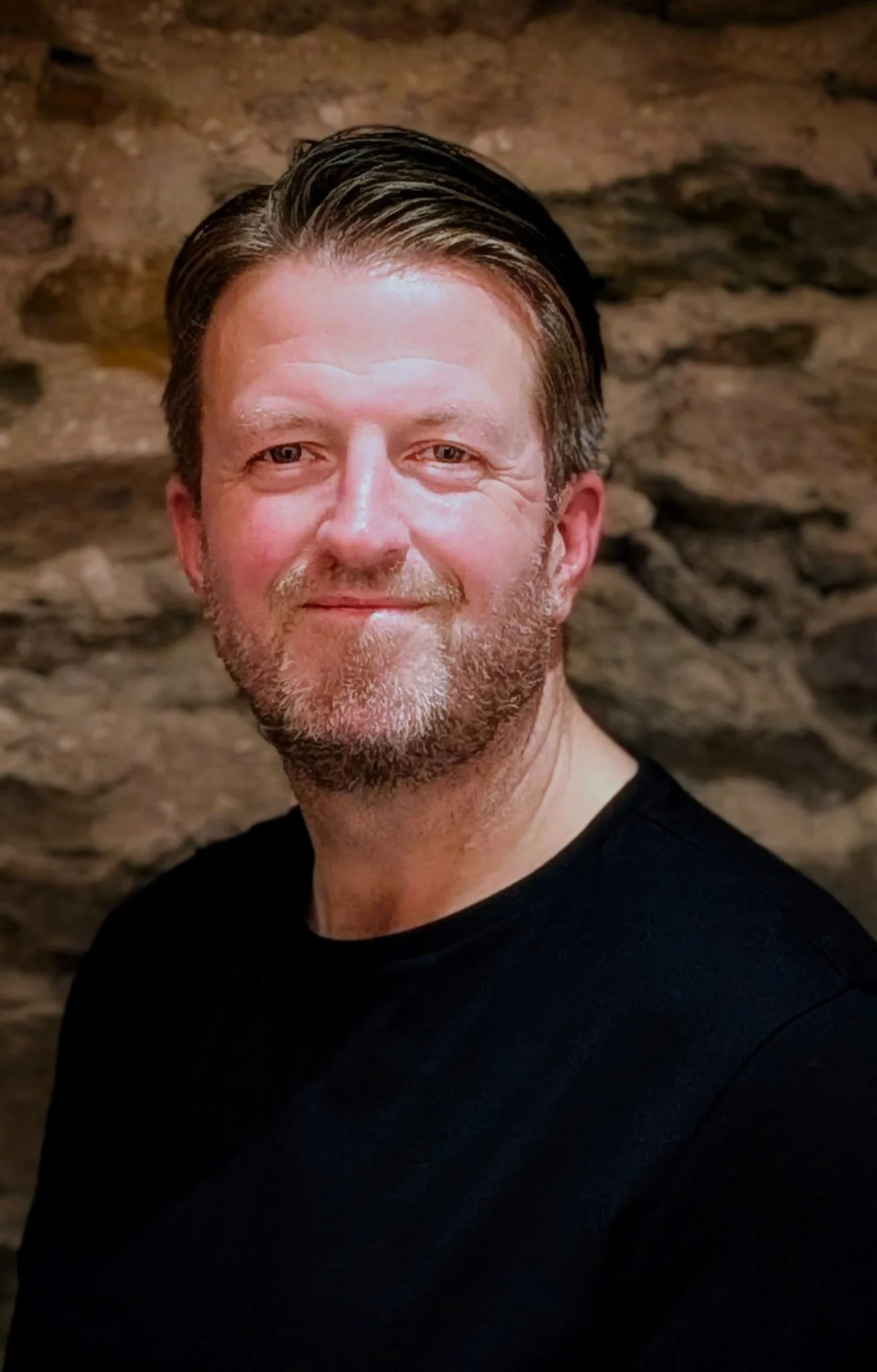 A middle-aged man with brown hair, a beard, and light skin, smiling, wearing a black shirt, standing in front of a stone wall.