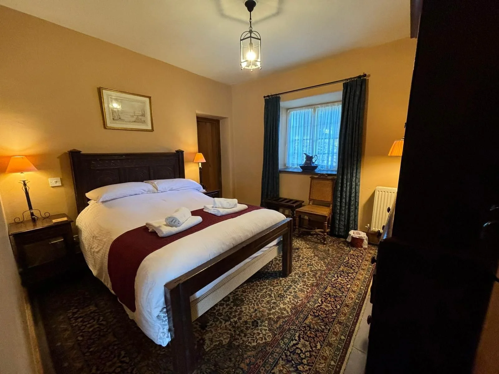 A cozy hotel bedroom with a large white bed, dark wood headboard, and matching side tables with lamps. There are two towels on the bed. A window with blue and black curtains, a chair, and a decorative item on the windowsill. The room has warm-colored walls, a patterned rug, and a light fixture hanging from the ceiling.