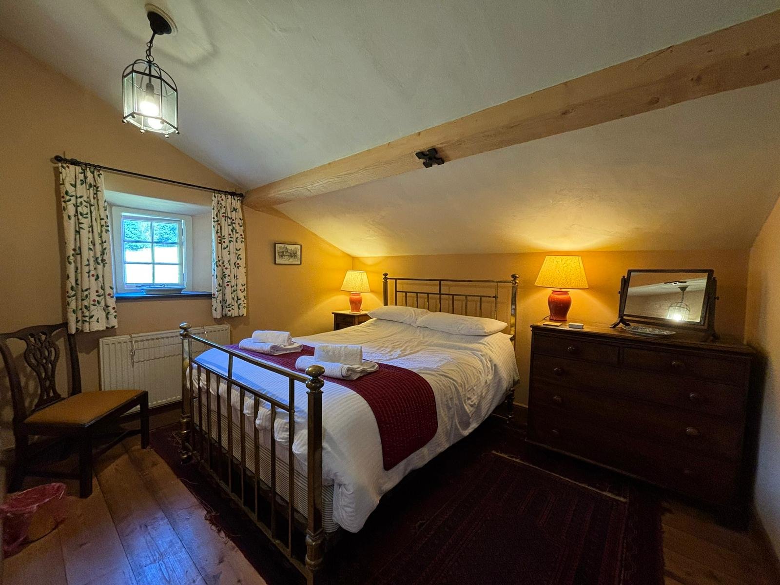 Cozy bedroom with a sloped ceiling, decorated with a metal bed, wooden dresser, mirror, lamps, window with floral curtains, and wooden chairs.