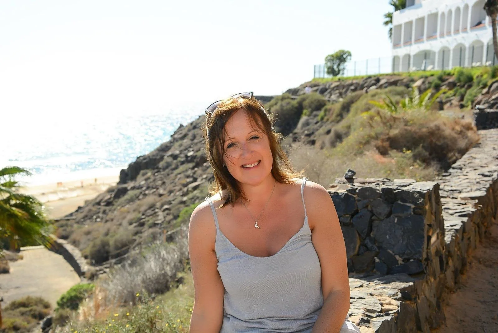 A woman with shoulder-length brown hair smiling in a sleeveless grey top sitting outdoors near rocky terrain and greenery, with the ocean and a modern white building in the background on a sunny day.