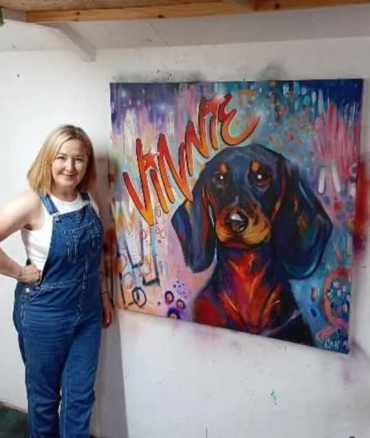 A woman standing next to a colorful painting of a dog with the name 'VINTIC' written on it, in an art studio or gallery setting.