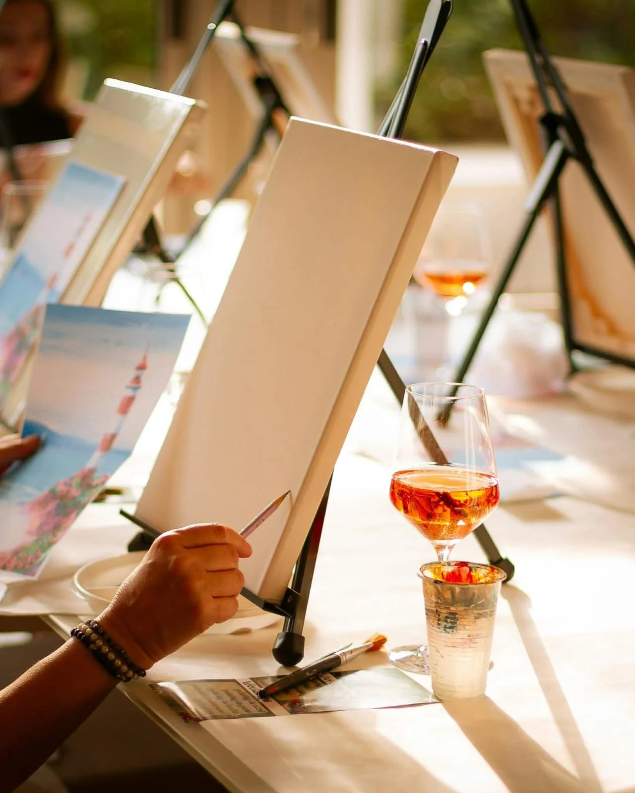An art class setup with blank canvases on easels, a person holding a paintbrush, and glasses of rosé wine on a table.