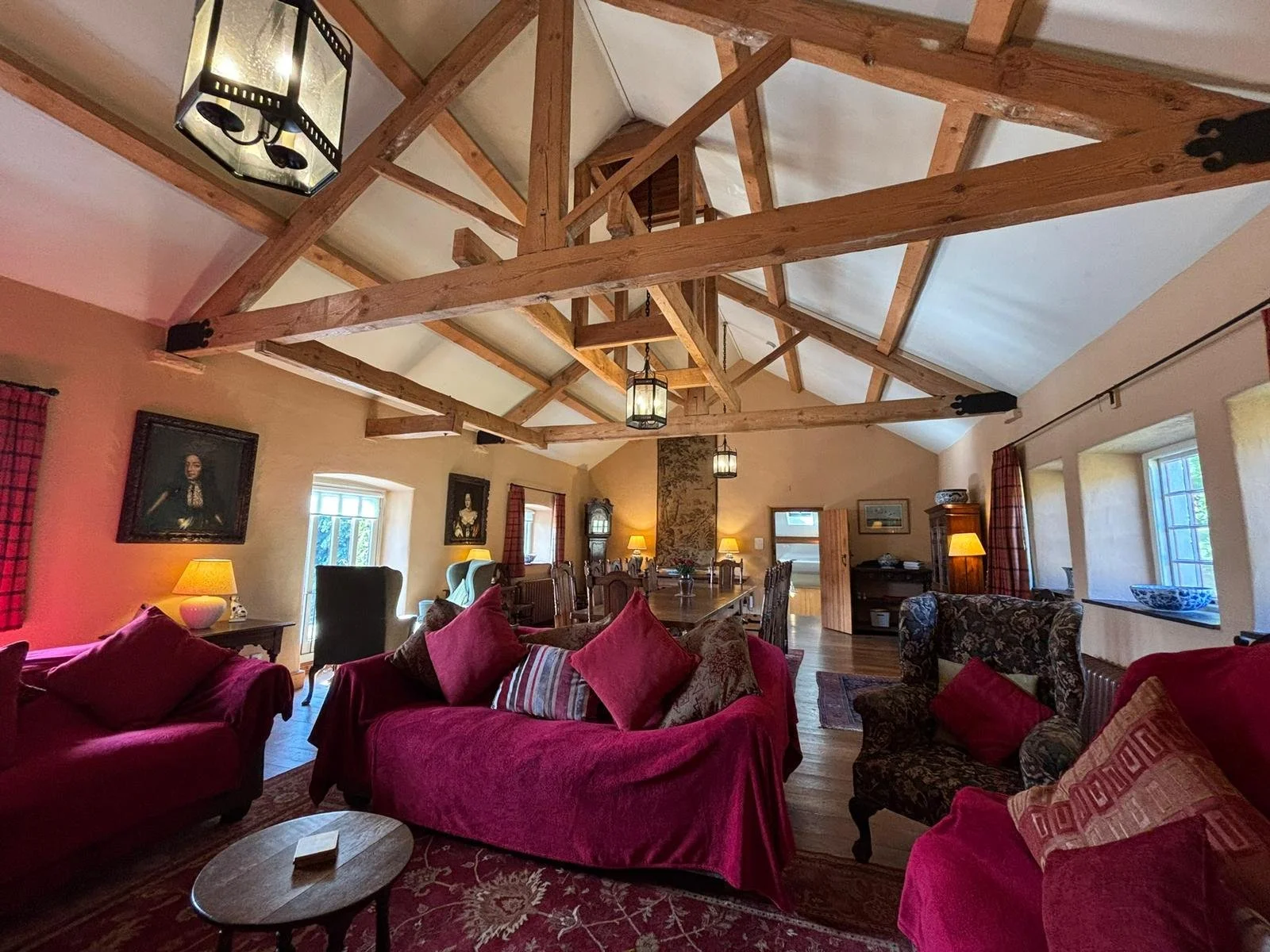 Living room with wooden ceiling beams, pink and patterned upholstered furniture, wooden table, lamps, paintings, and windows with red curtains.