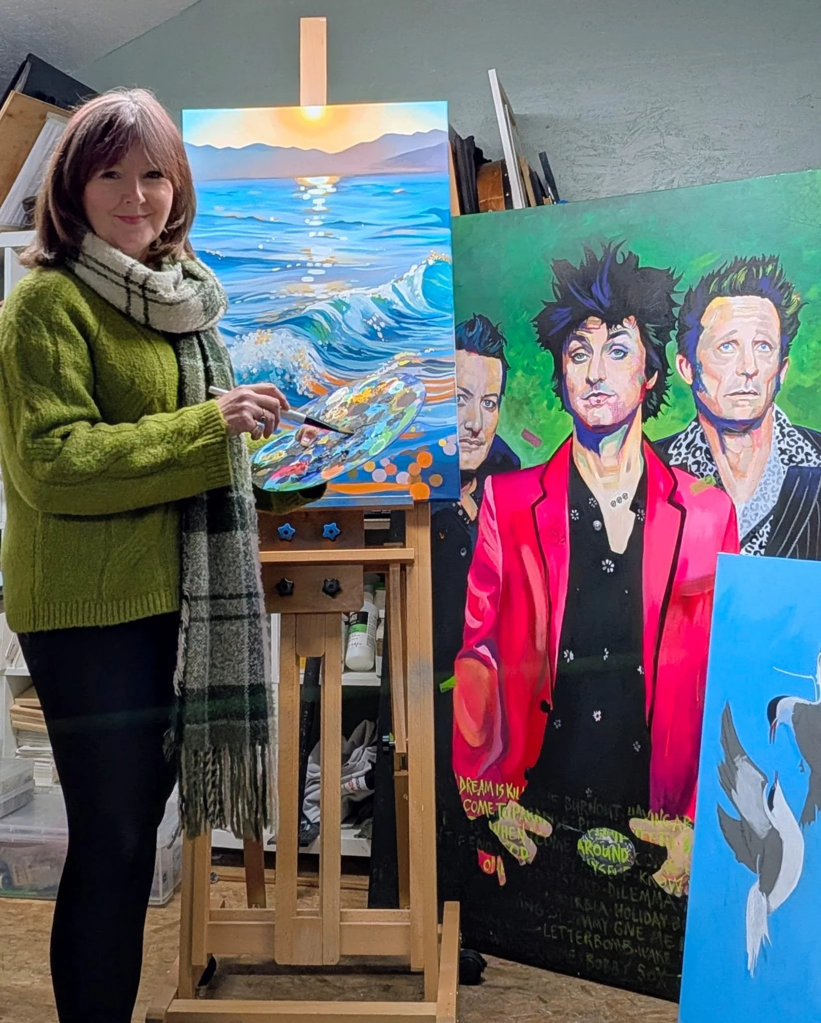 An woman standing in an art studio holding a paint palette and brush, with colorful paintings of ocean waves and portrait of three women on easels