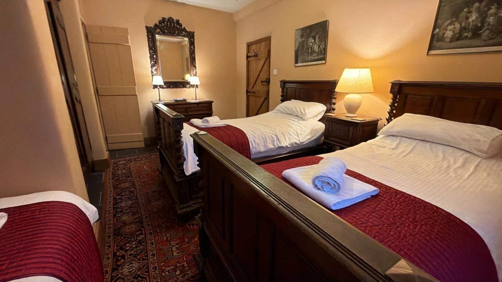 A cozy bedroom with three beds, each with white linens and red bed runners, wooden furniture including headboards, bedside tables, a dresser with a mirror, and a lamp, with framed artwork on the walls.