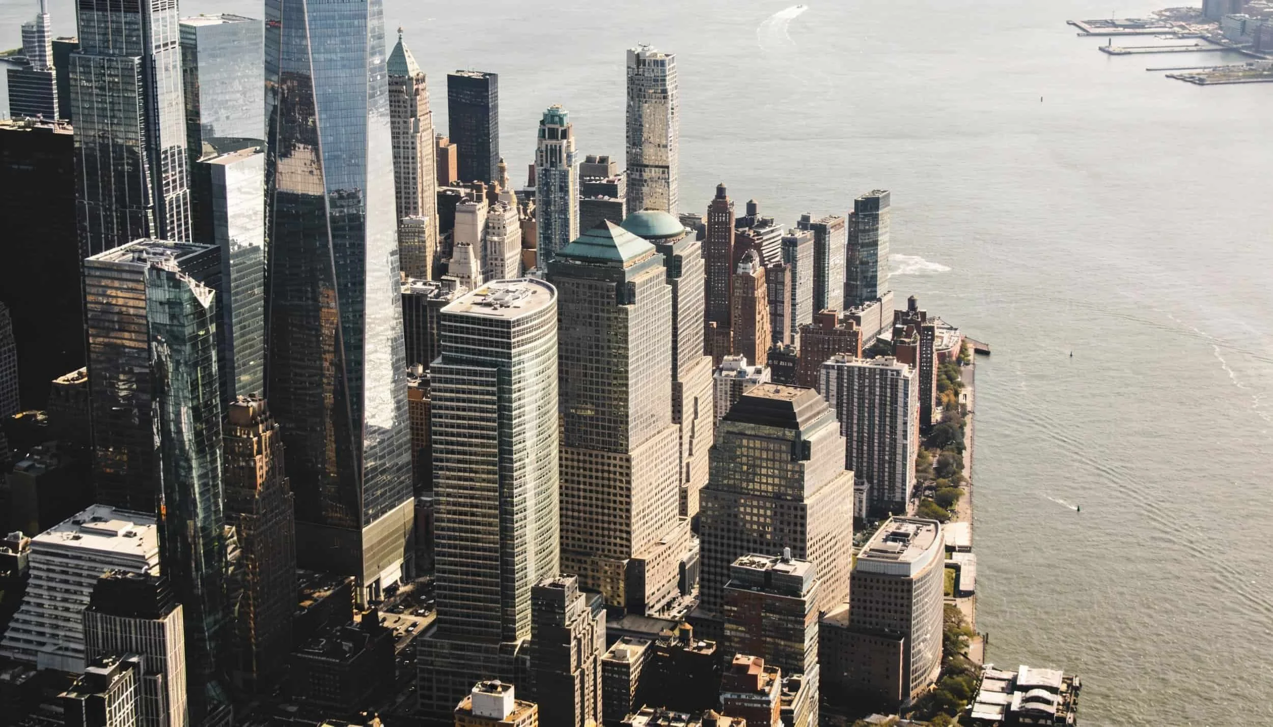 Aerial view of a city skyline with tall modern skyscrapers along a body of water, featuring glass facades reflecting sunlight.