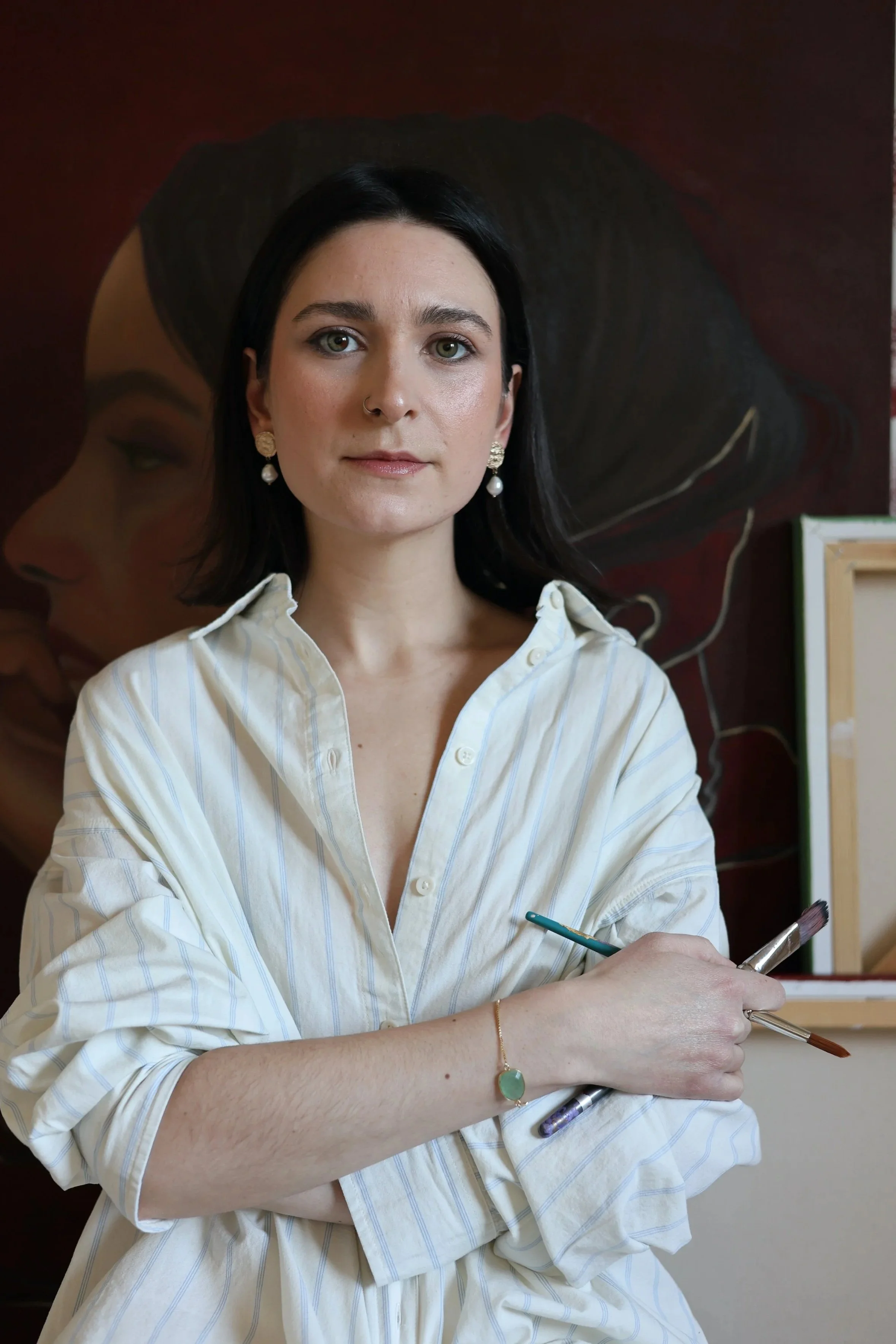 A female artist with black hair, wearing a white striped shirt and earrings, holding paintbrushes, standing in front of a large painting of a woman's profile with dark hair holding a burning candle.