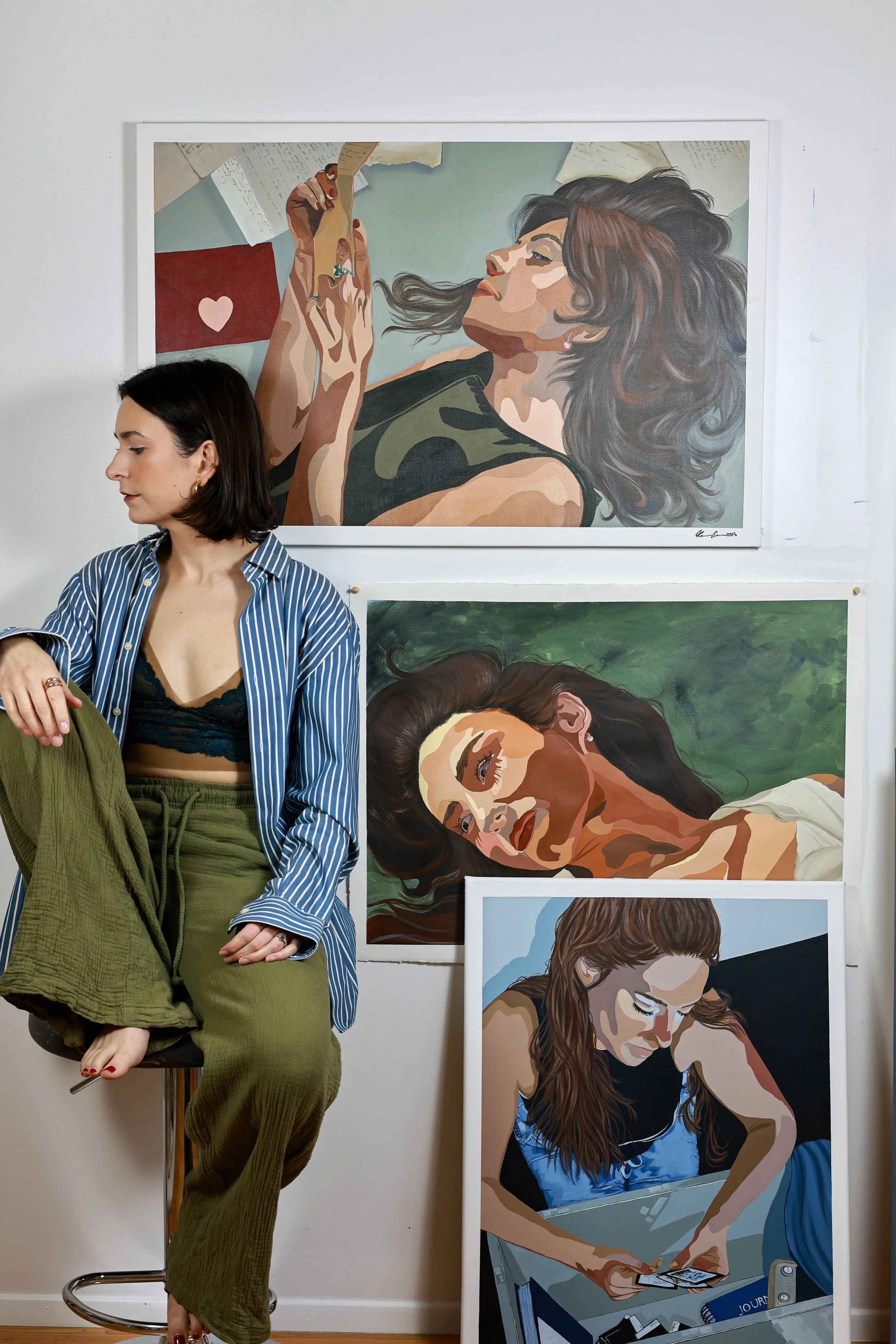 A woman with dark hair and a striped blue and white shirt sitting on a stool in front of three artwork pieces of women—two are lying down and one is reading a letter—displayed on a white gallery wall.