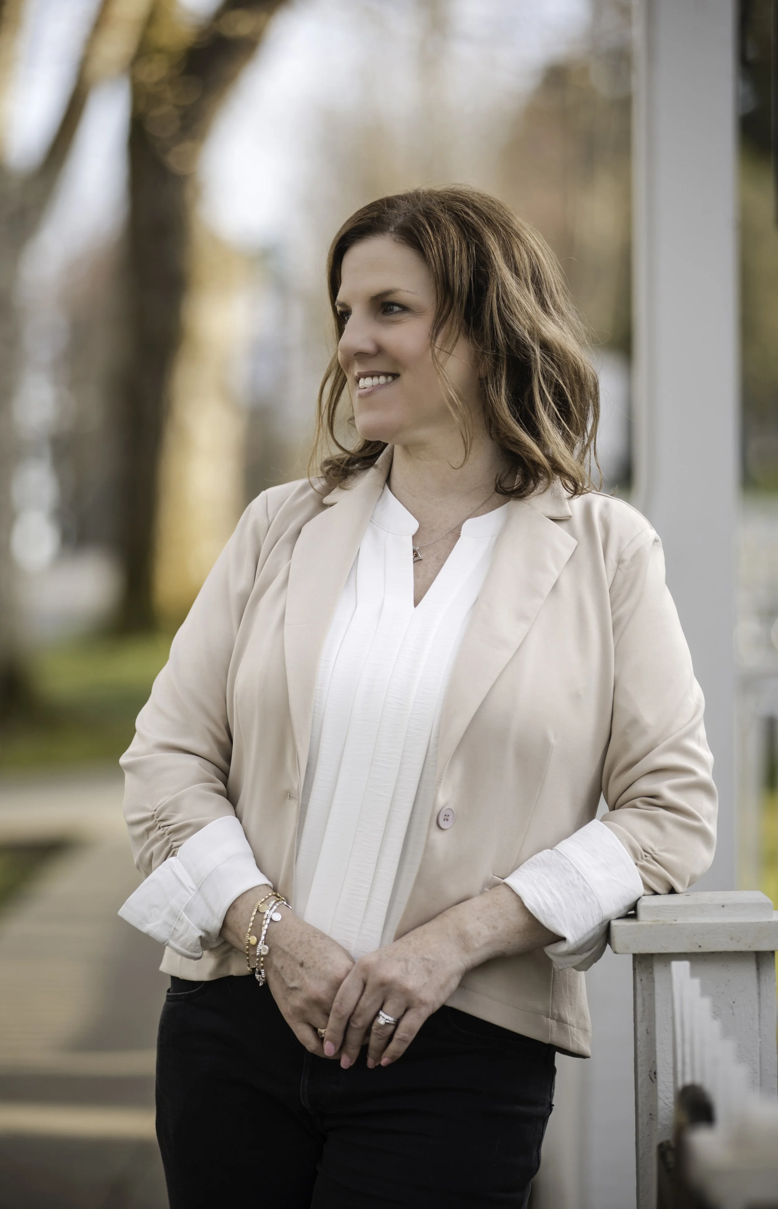 A woman with shoulder-length brown hair wearing a beige blazer and white blouse, standing outdoors with a blurred background of trees and sunlight, smiling and looking to her left.