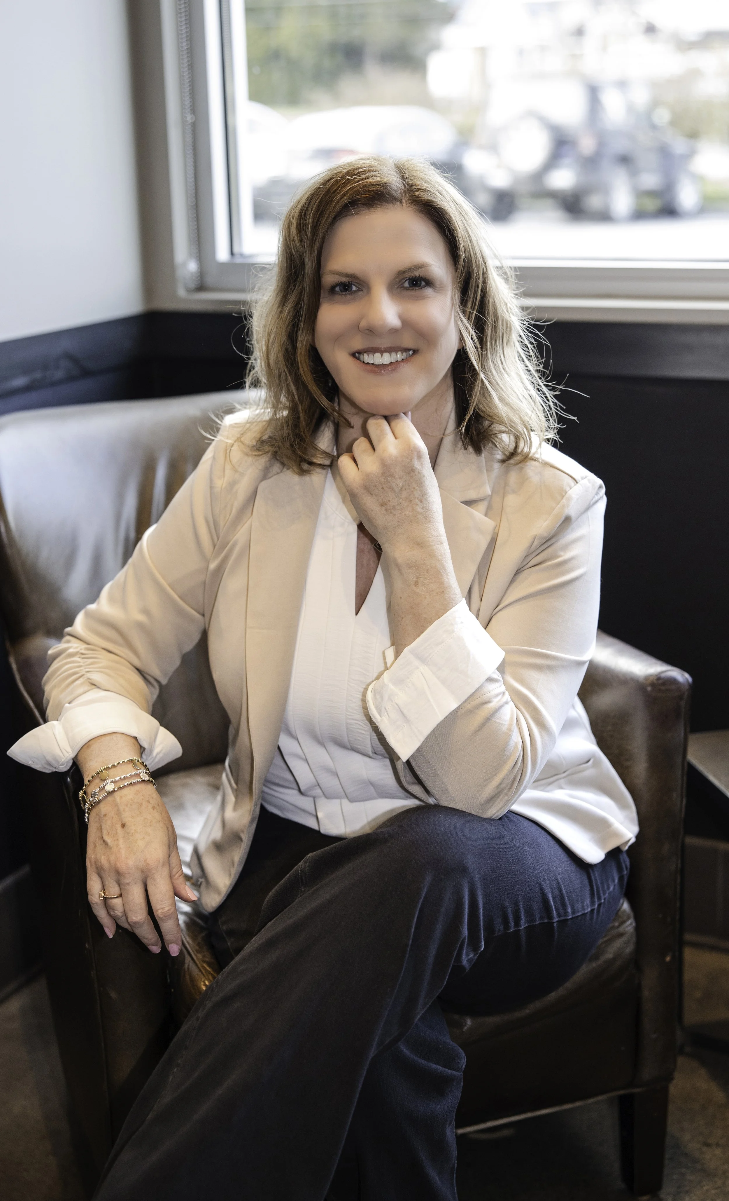A woman with shoulder-length wavy hair, wearing a cream blazer, white blouse, and dark pants, sitting on a brown leather chair near a window, smiling and resting her chin on her hand.