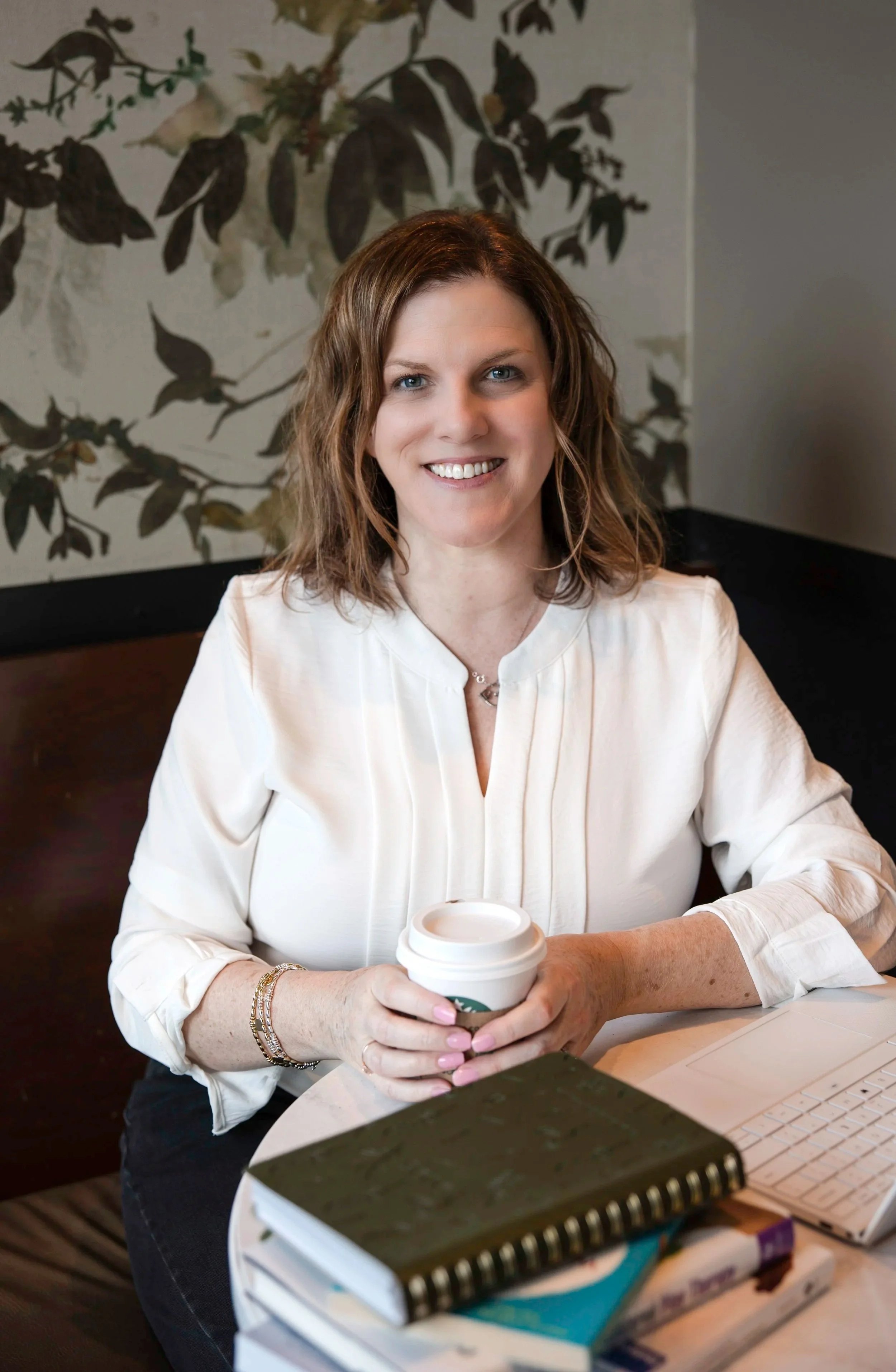 A woman with shoulder-length wavy brown hair, wearing a white blouse, sitting at a table in a cafe. She is smiling, holding a to-go coffee cup. There are notebooks and a computer keyboard on the table.