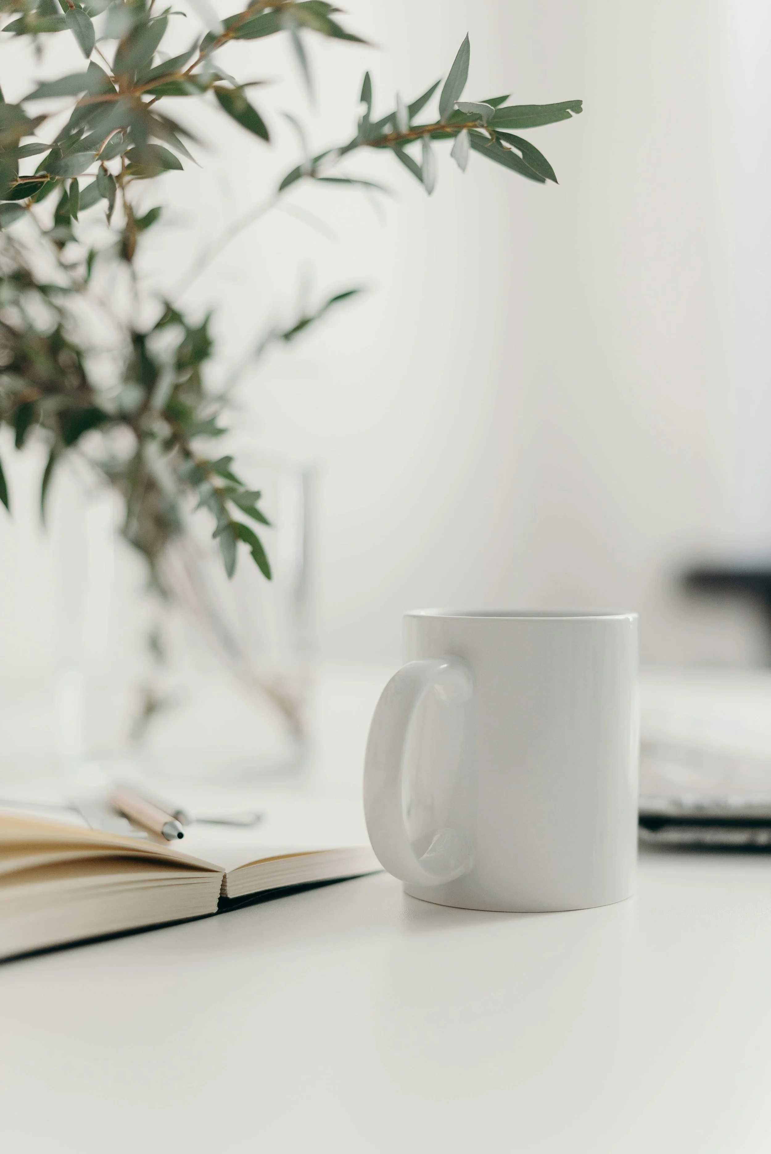 A white ceramic mug, an open notebook with a pen on top, and a potted plant are arranged on a white desk in a bright, minimalistic setting.