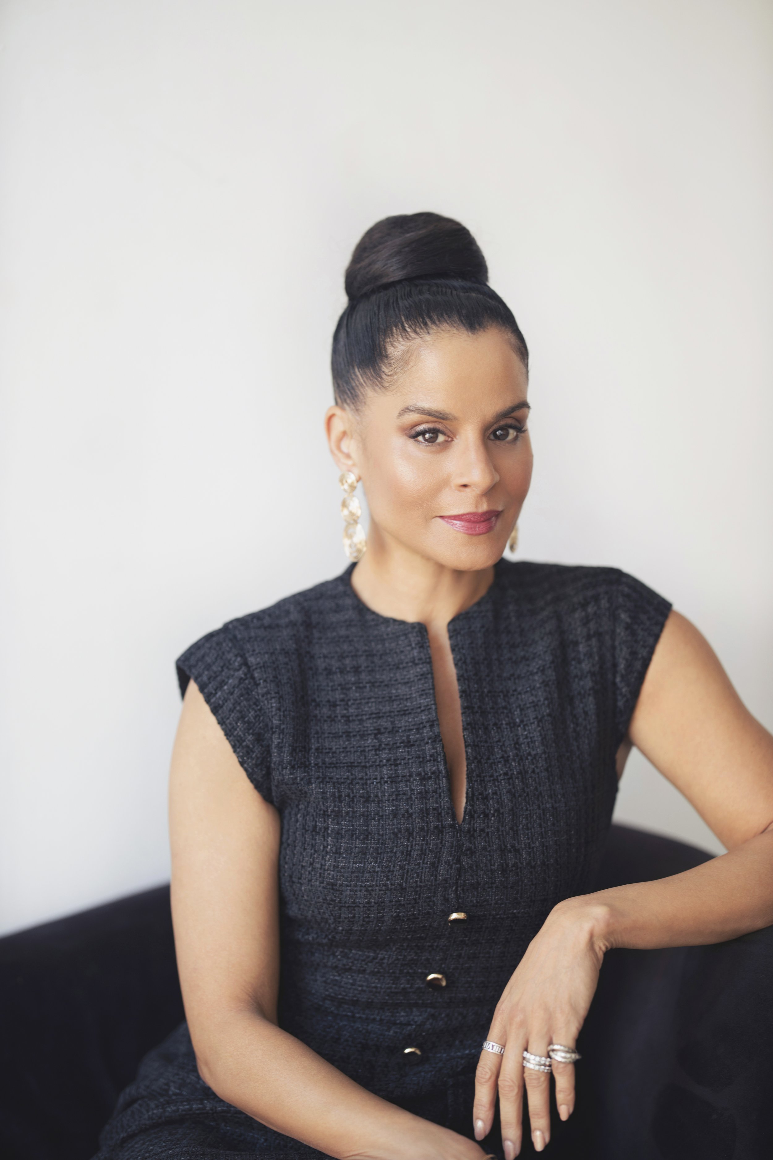 A woman with dark hair in an elegant updo, wearing a black textured sleeveless dress and gold earrings, sitting on a black sofa against a plain light background.