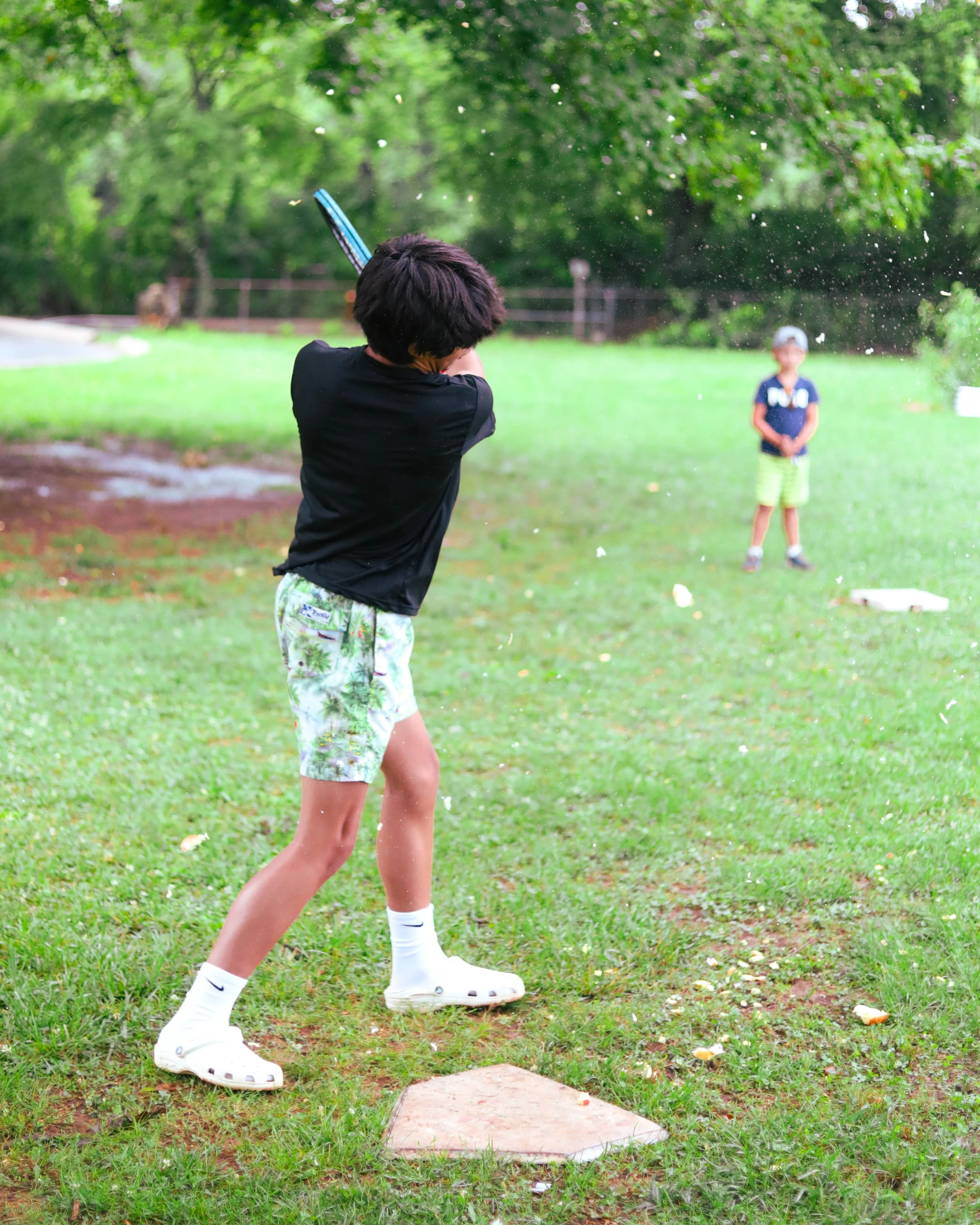Two children playing catch outside on a grassy field; one is swinging a bat, and the other stands in the background.