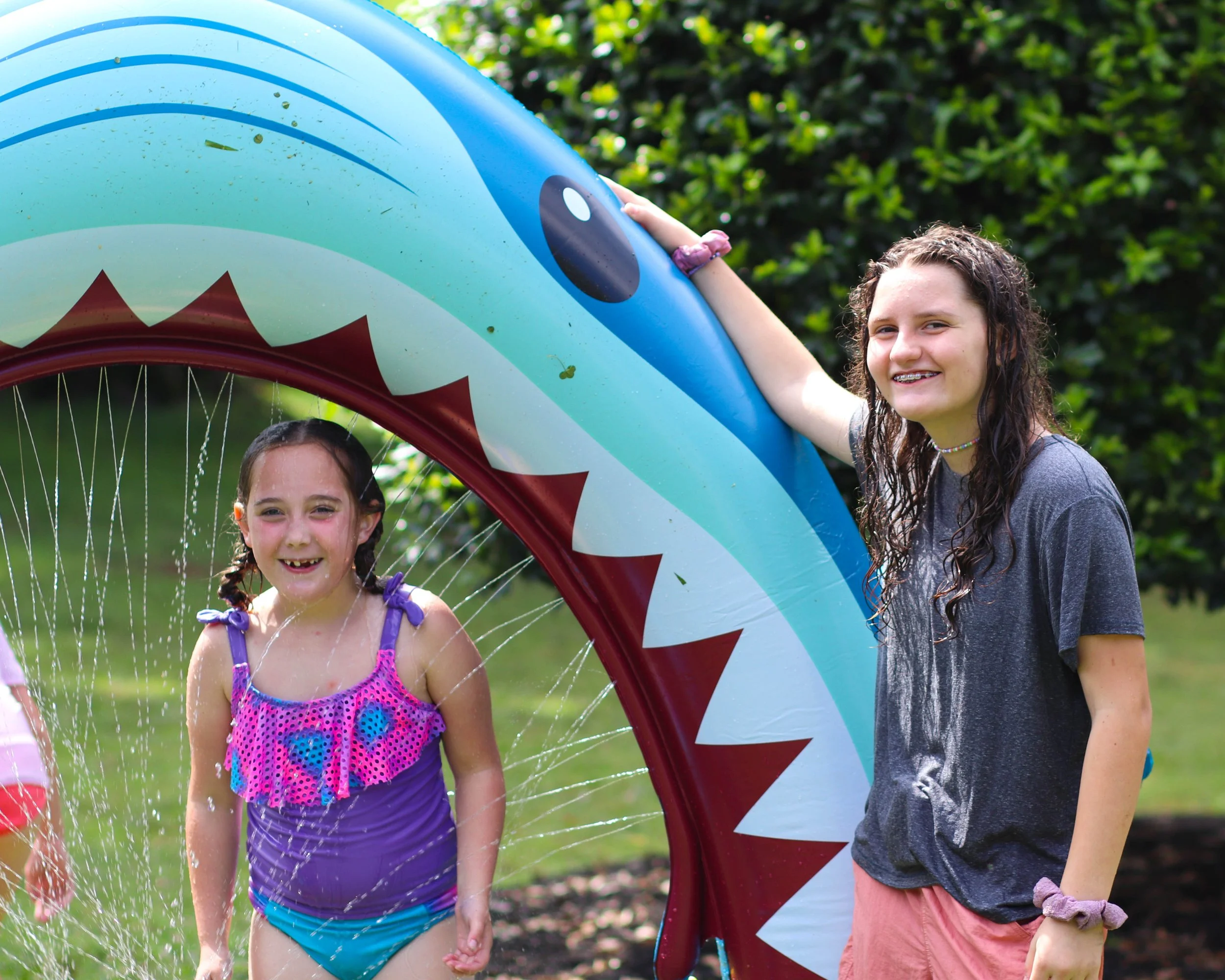 Two girls playing outdoors with inflatable shark-shaped water slide or pool, one girl is inside getting wet, and the other girl is outside smiling, with green foliage in the background.